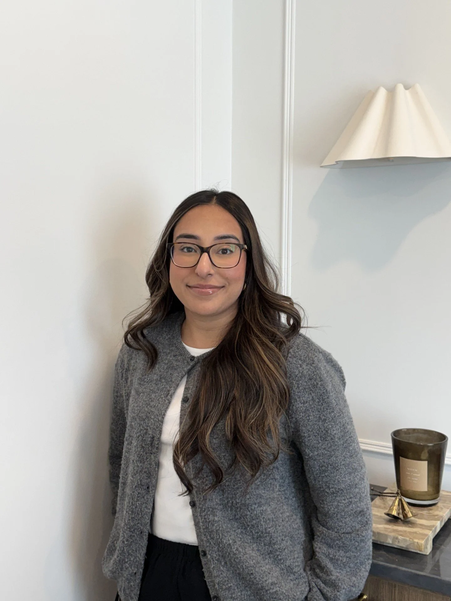A woman with brown hair wearing a grey shirt standing near a wooden cabinet with a lamp on the wall behind her.