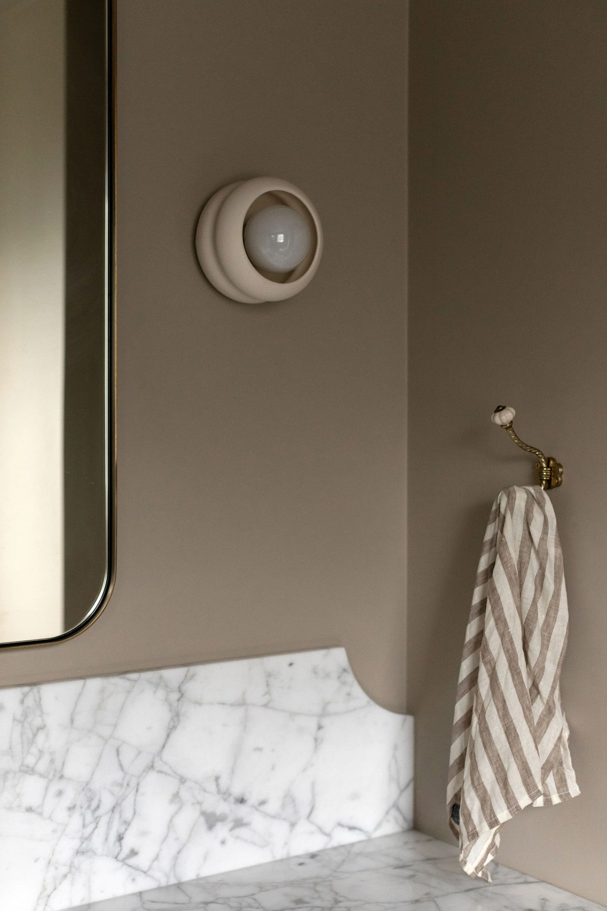 Bathroom detail featuring a sculptural wall sconce, marble backsplash and brass towel hook against warm neutral walls.