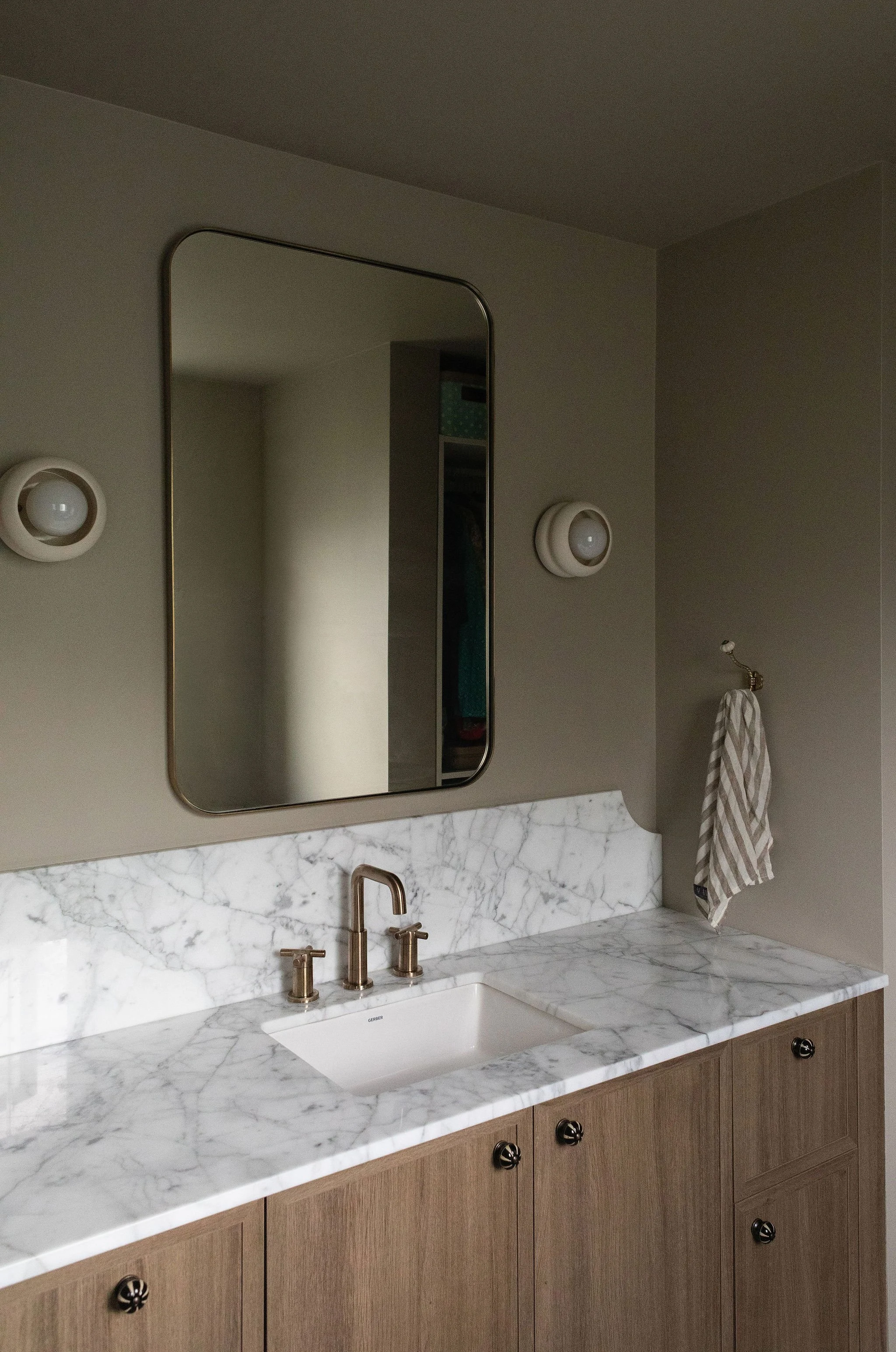 Bathroom vanity with white marble countertops, a natural wood cabinet, brass faucets and a rounded rectangular mirror.