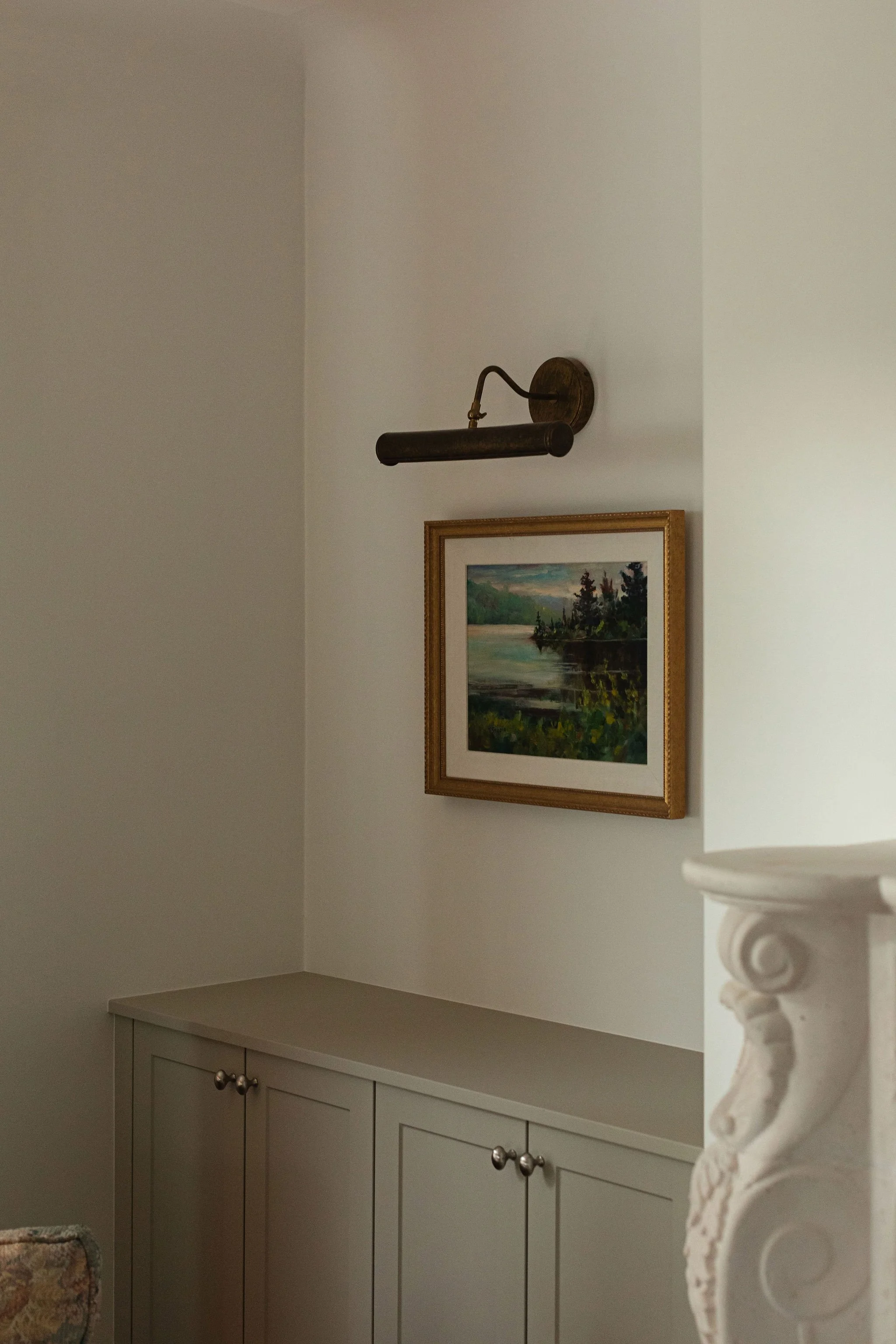 Custom taupe living room cabinetry with brass picture lighting and framed landscape artwork.