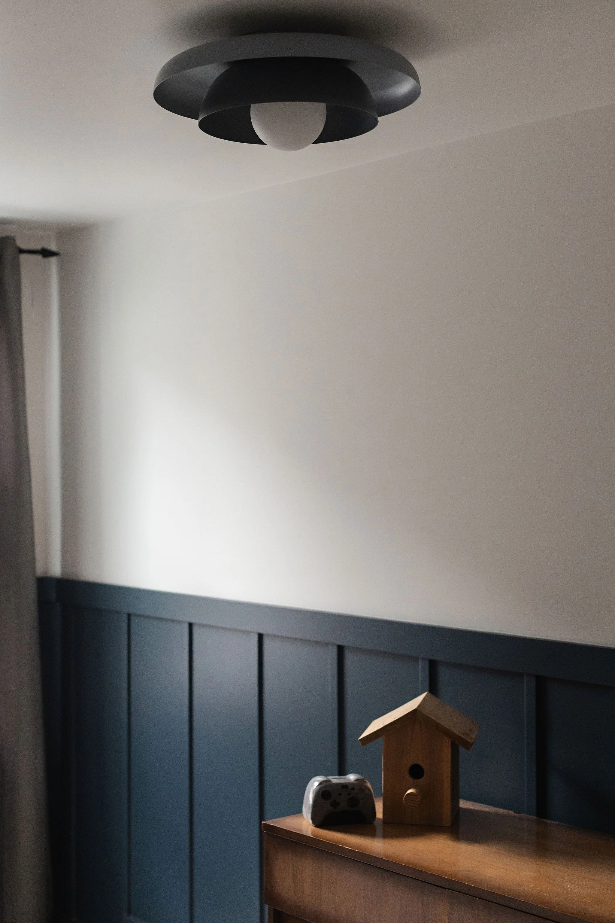 Bedroom with deep blue-green wall panelling, a warm wood dresser and a simple black flush-mount ceiling light.