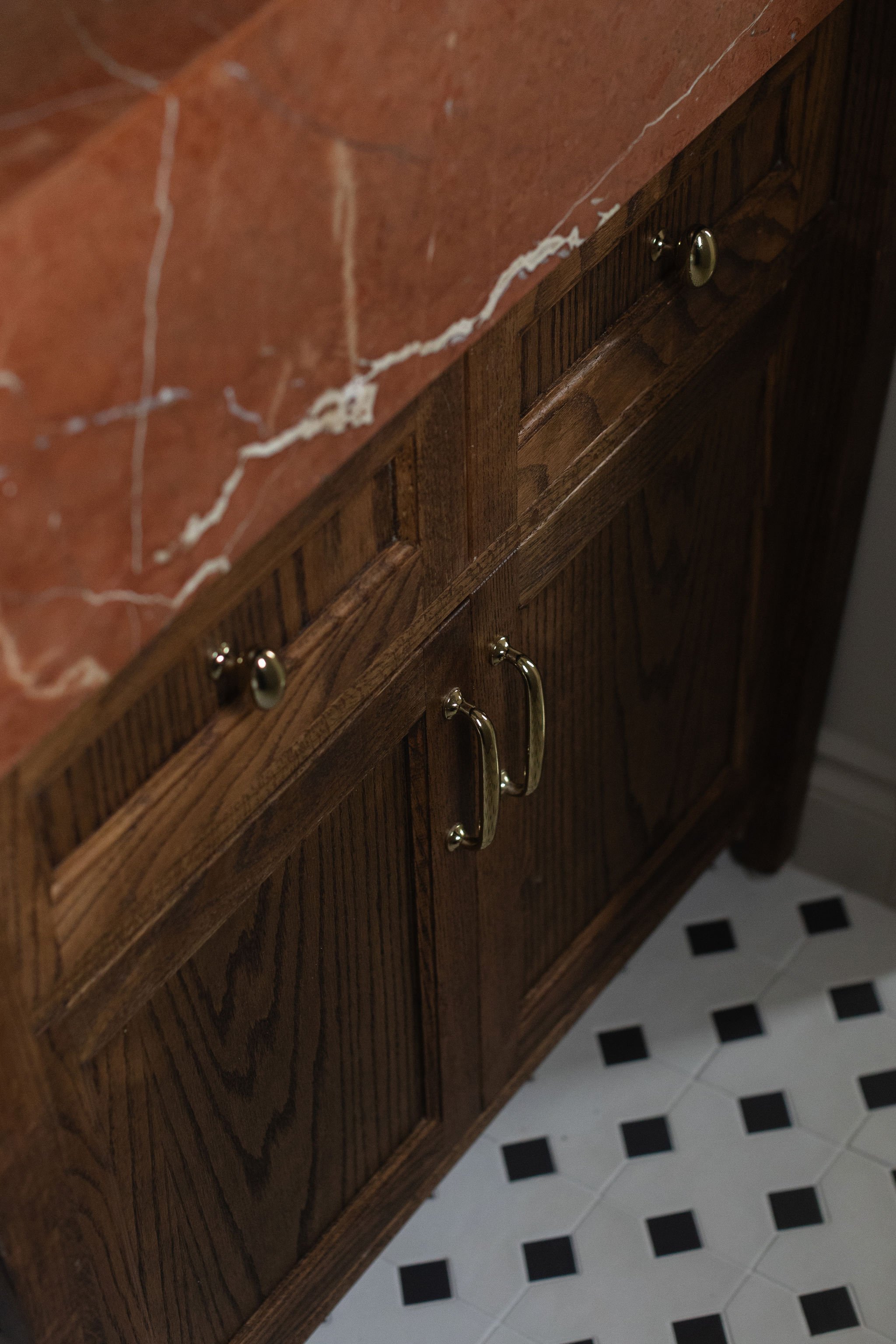 Dark wood bathroom vanity with traditional brass hardware, red marble countertop and black and white patterned floor tile.