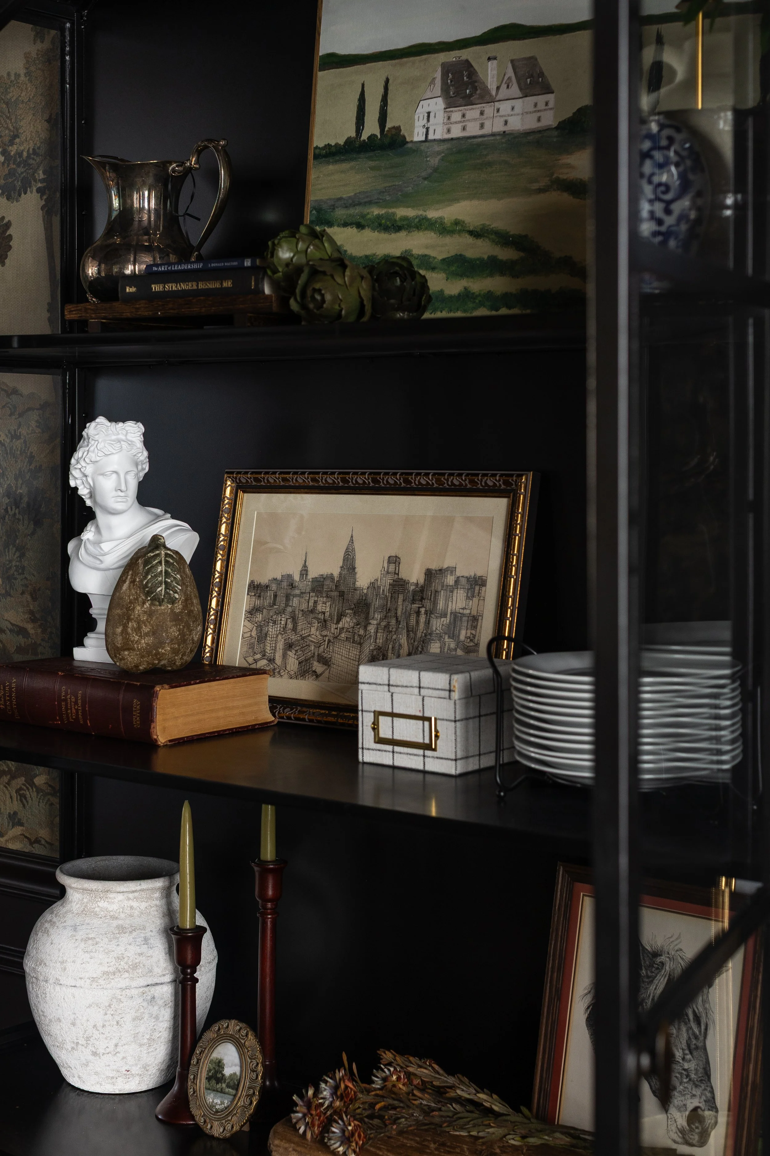 Black display cabinet with decorative items including a silver pitcher, art books, art prints, a bust sculpture, artichokes, a stone, a gray box, white plates, a large white vase, candlesticks, and framed artwork.