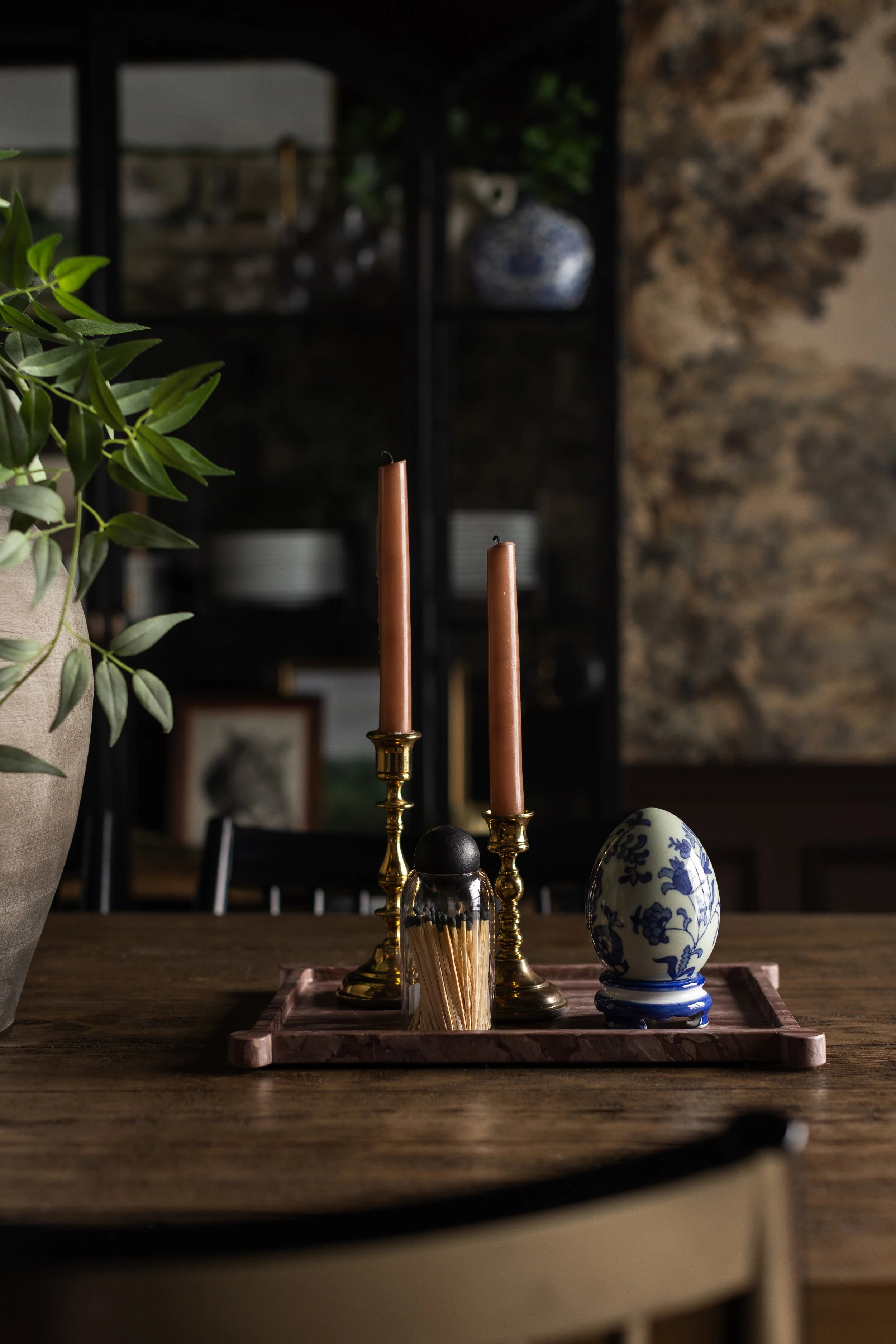 Decorative table setting with two pink candles in gold holders, a blue and white floral egg, a matchstick holder, and green foliage on a wooden table.