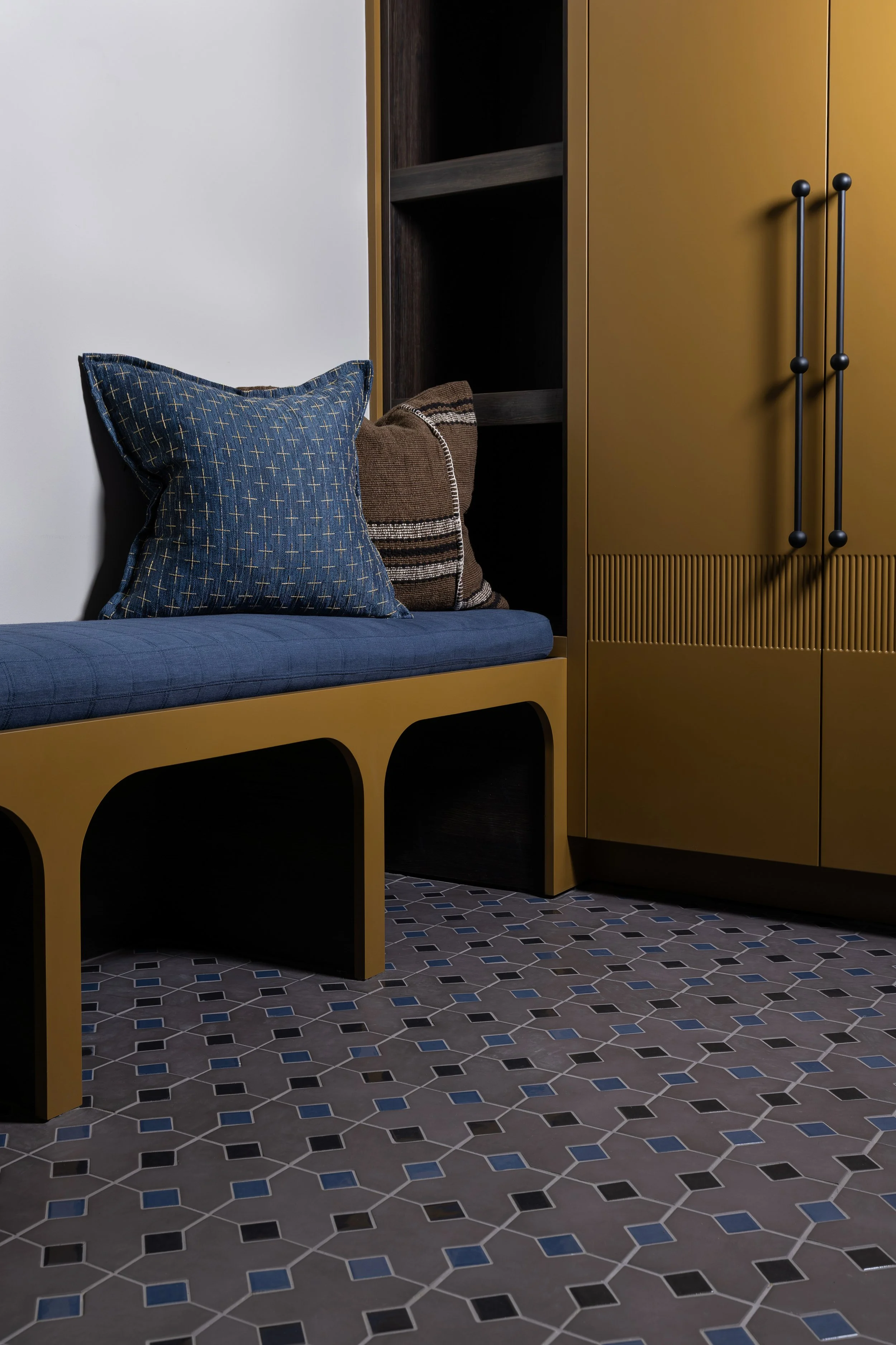 Indoor space with mustard-colored storage cabinet, black shelving, a blue cushioned bench, and decorative pillows, set on a patterned tile floor.
