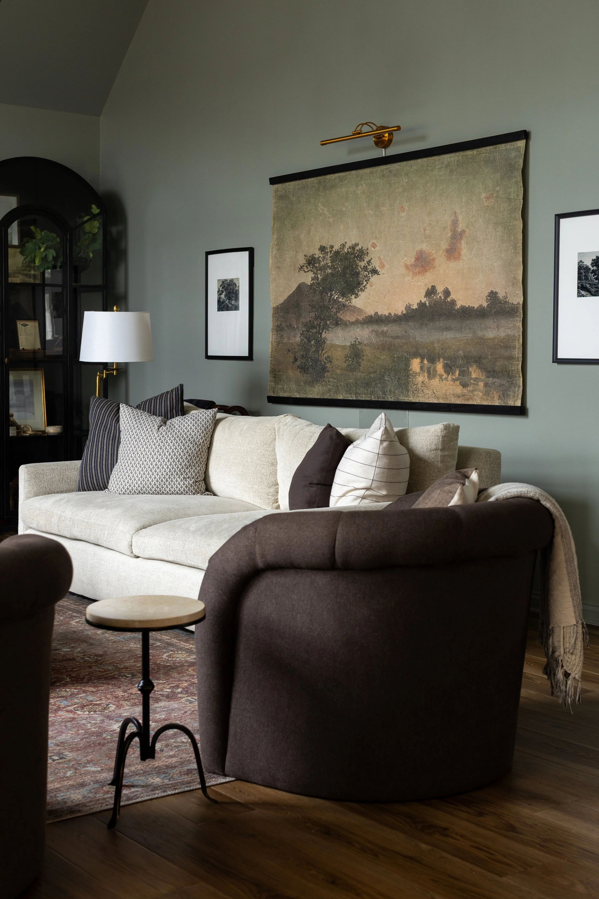 A cozy living room with a beige sofa adorned with various pillows, a dark armchair, a small round side table, a patterned rug, framed black-and-white photos on a sage green wall, a vintage landscape tapestry, a white floor lamp, and a black display c