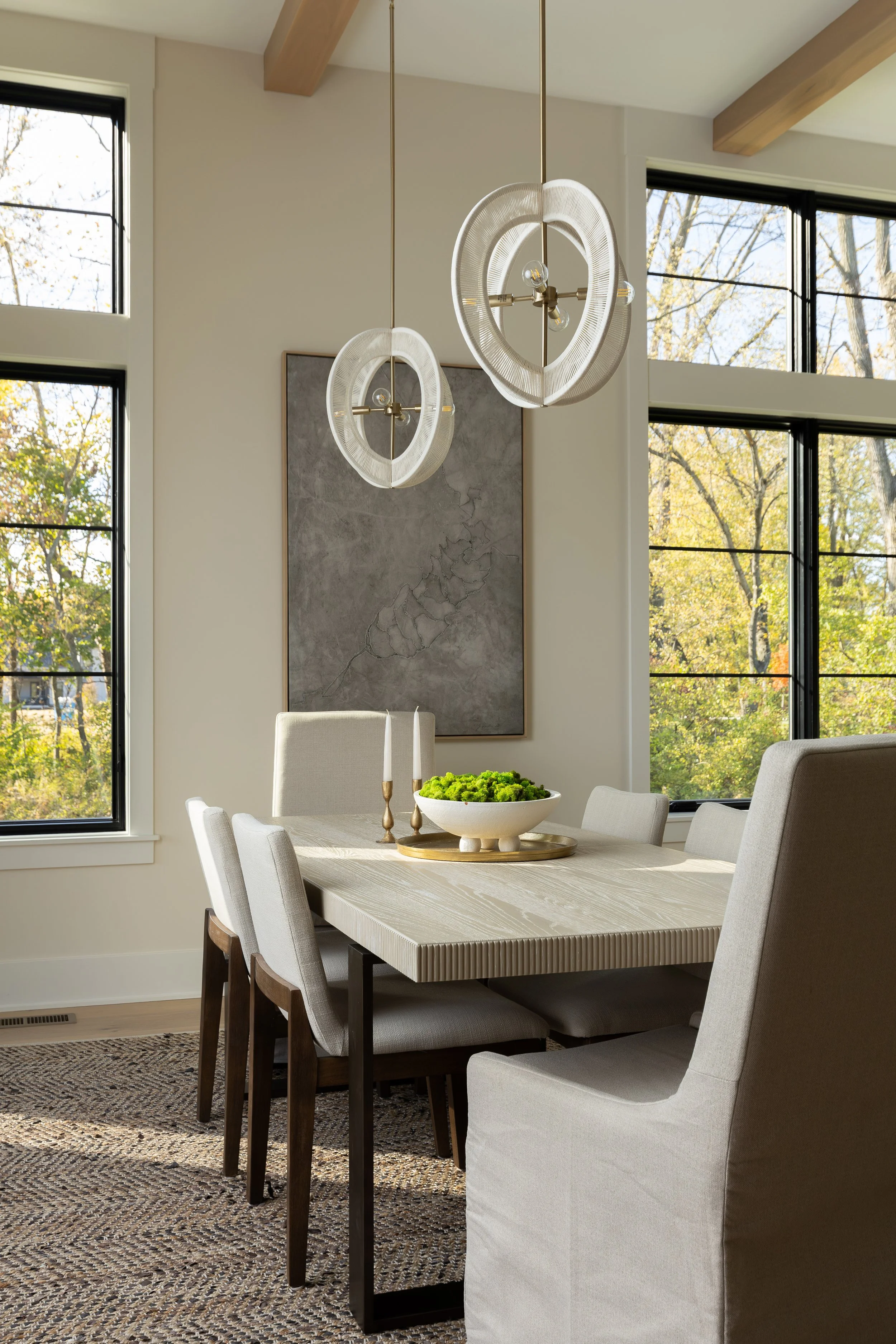 A modern dining room with a light wooden table, six upholstered chairs, large windows with black frames showing an outdoor view of trees, two contemporary pendant light fixtures hanging from the ceiling, a textured rug, a decorative bowl with green m