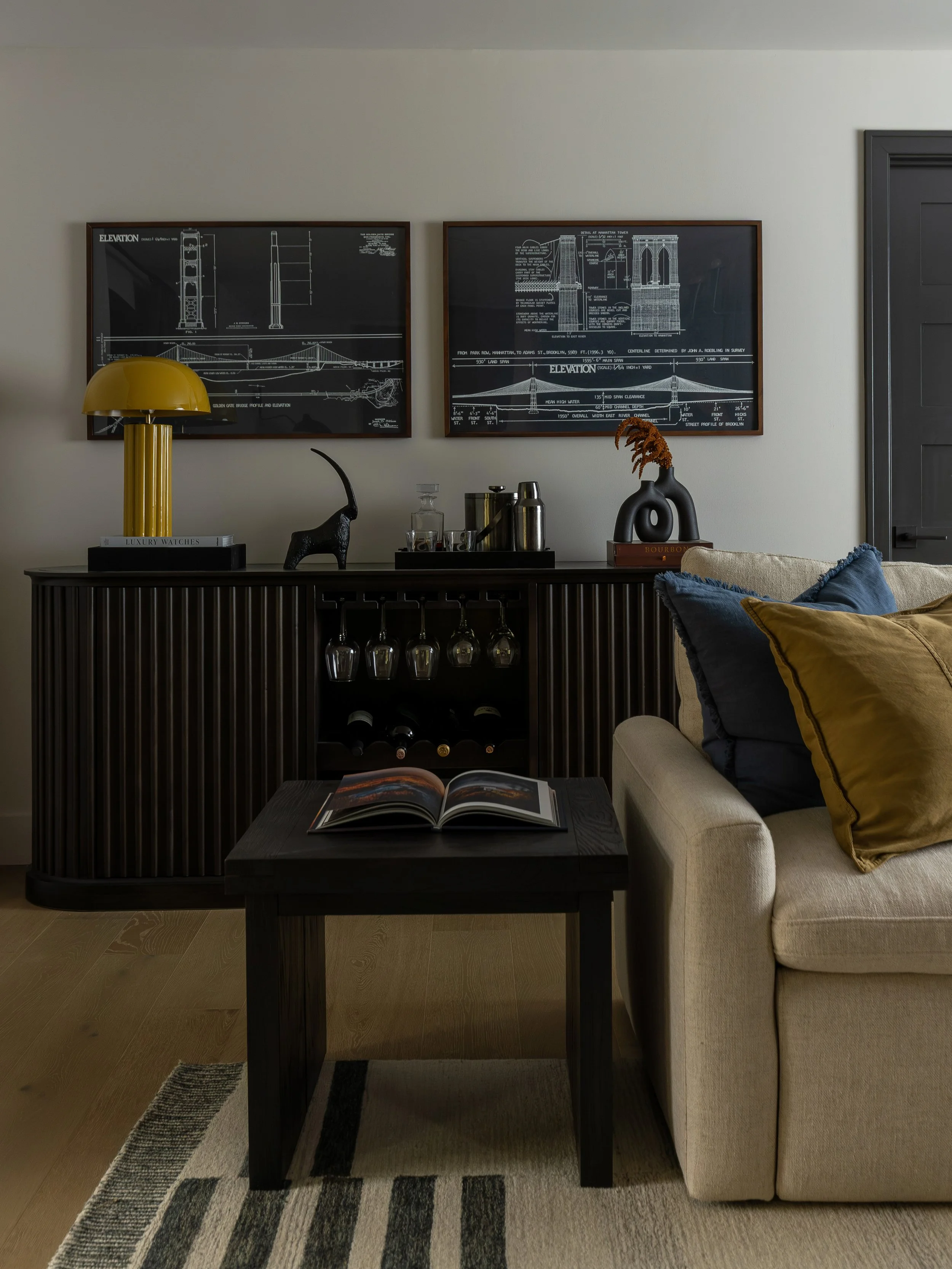Living room with black sideboard, wall art of architectural blueprints, beige sofa with blue and mustard pillows, and a black coffee table with an open book. Decor includes a yellow lamp, black sculpture, glass decanter, and a vase with dried orange 