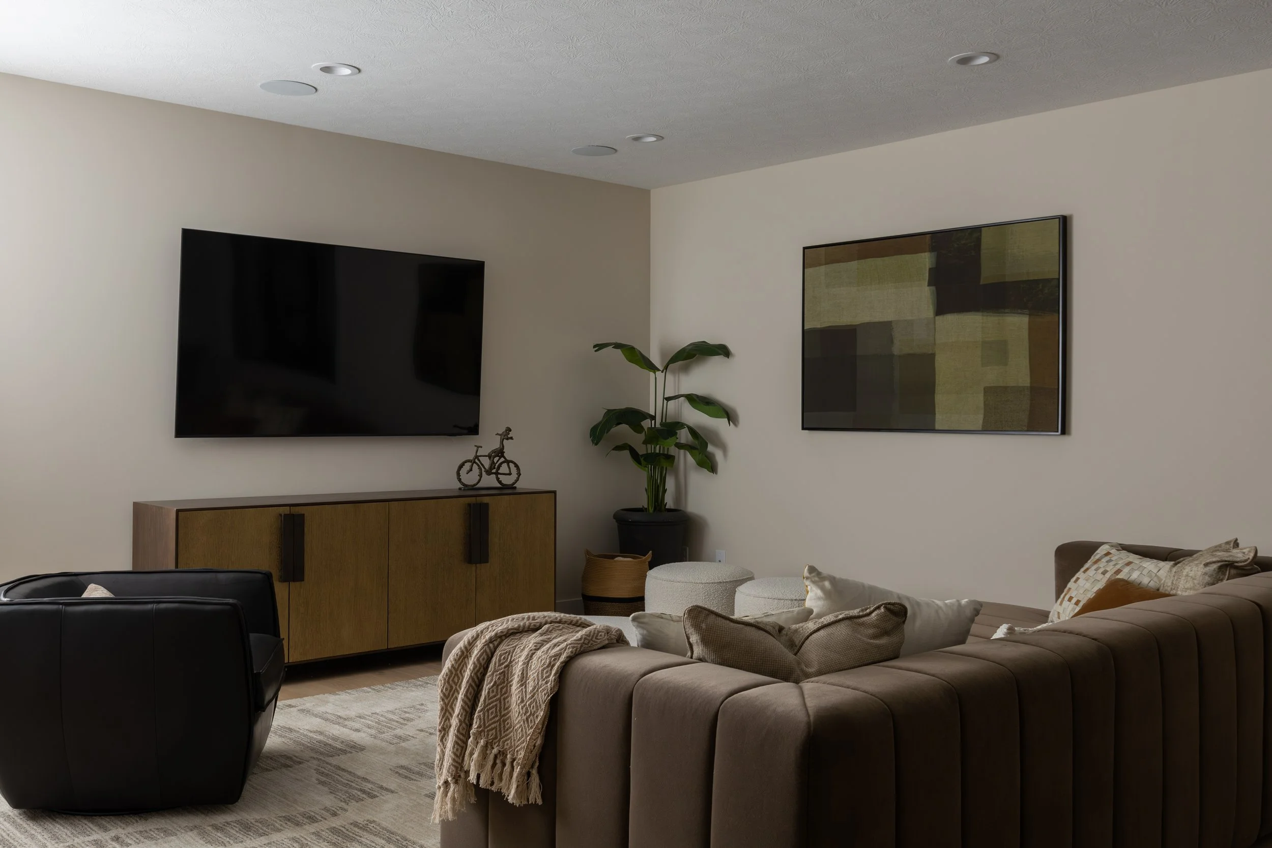 Living room with a large beige sectional sofa, a black armchair, wall-mounted TV, wooden cabinet, potted plant, framed artwork, and decorative pillows and throw blankets.