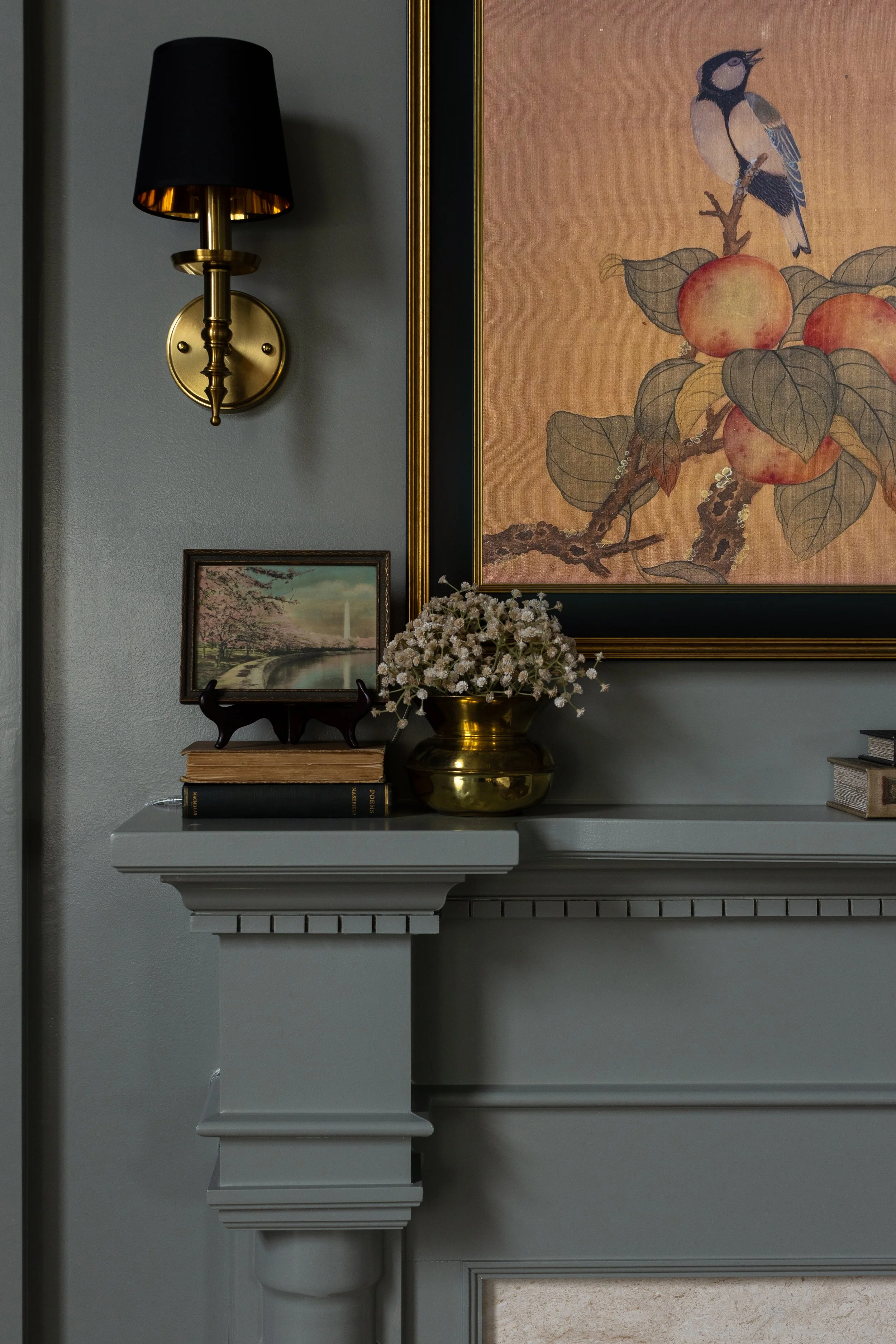 A living room corner with a gray fireplace mantel, a black and gold wall sconce, a framed landscape painting, a small bouquet of white flowers in a gold vase, and a stack of books.