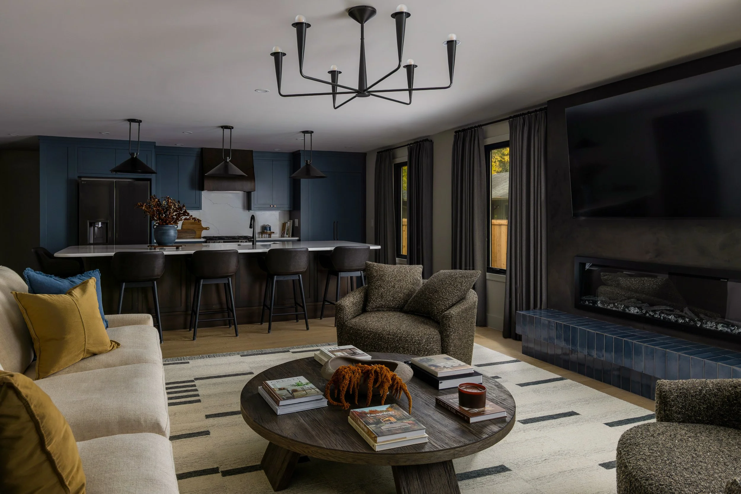 Living room with beige sofa, brown armchairs, coffee table with books and decor, black ceiling light, dark curtains, fireplace with blue tiles, flat-screen TV, open kitchen with island, black chairs, and pendant lights.