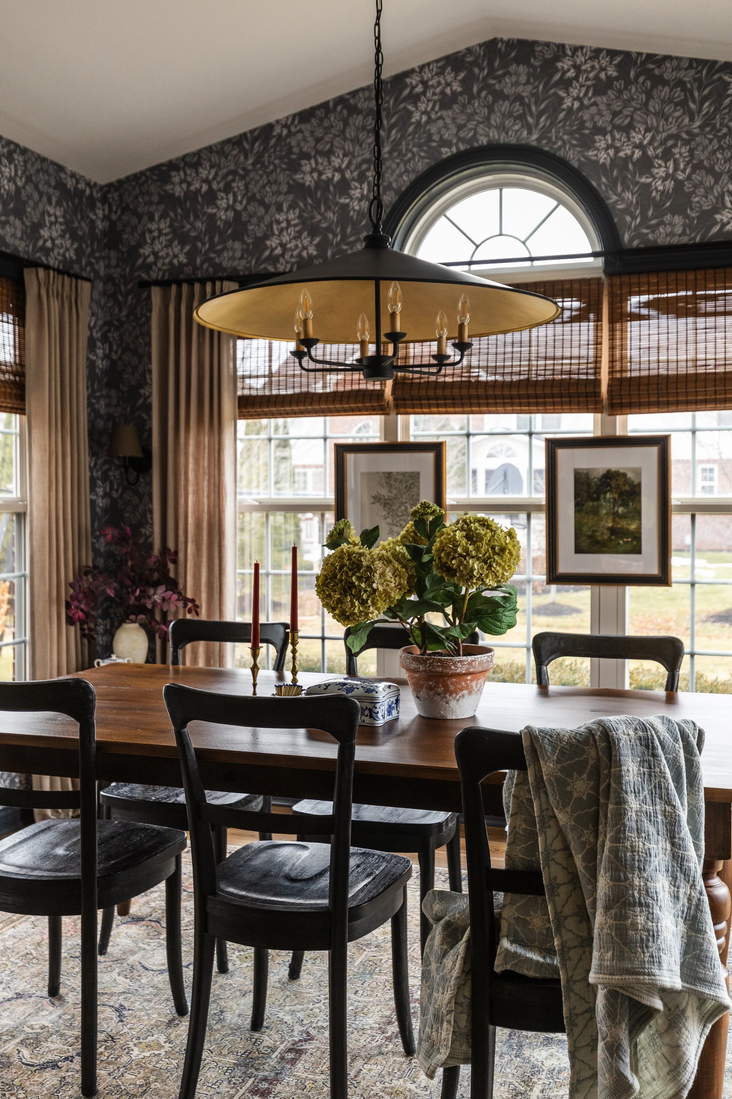 A dining room with a wooden table, surrounded by black chairs, a large floral centerpiece, framed pictures on the window, a hanging chandelier, and patterned wallpaper.