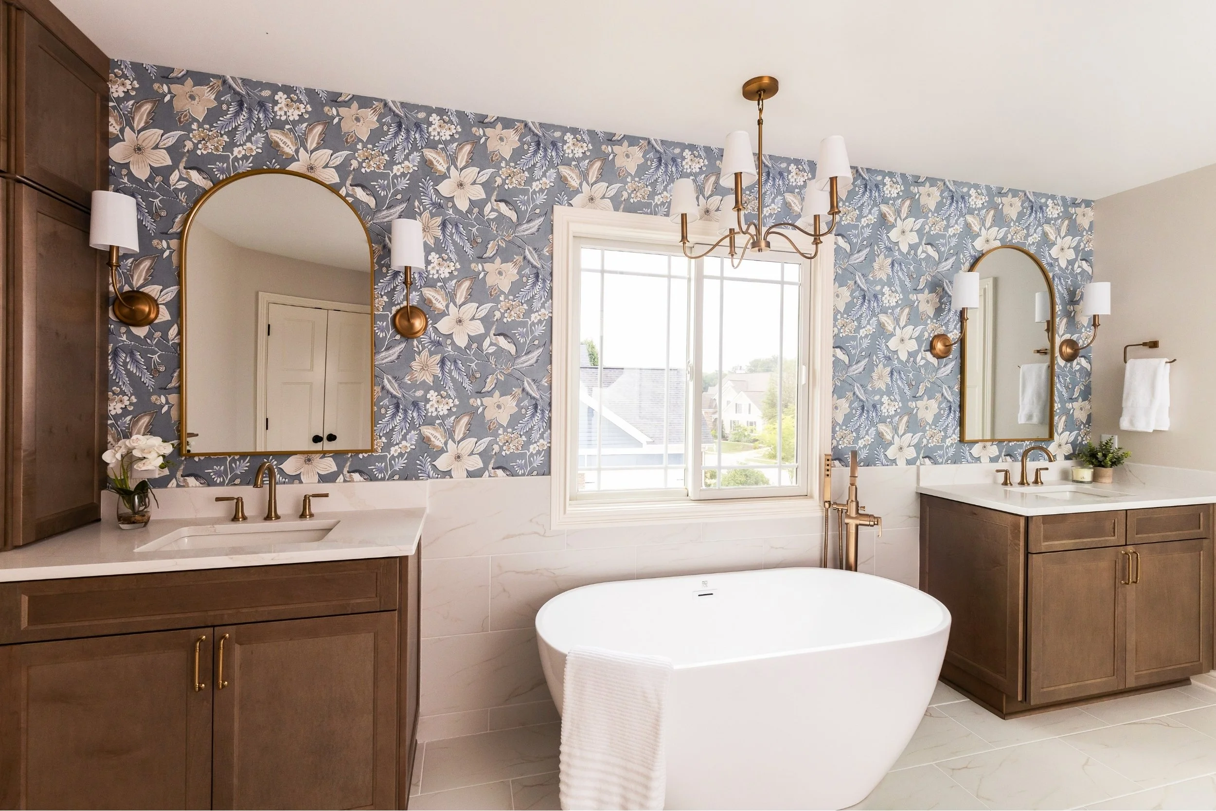 Bathroom with a freestanding bathtub, two wooden vanities with white countertops, arched mirrors, floral wallpaper, window, chandelier, and wall sconces.