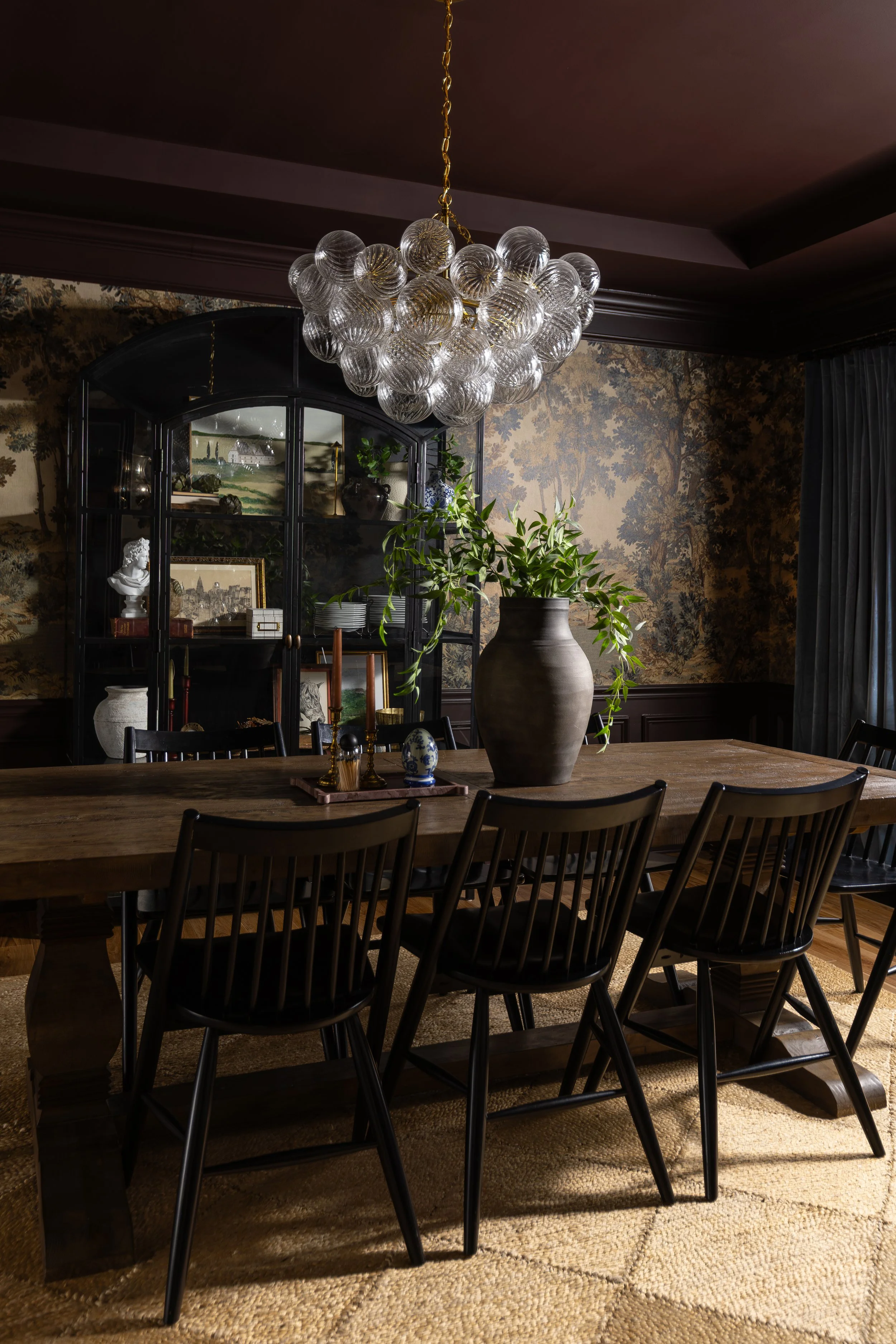 A dining room with a large wooden table with black chairs, a large vase with green plants, a black display cabinet, and a modern chandelier hanging from the ceiling.