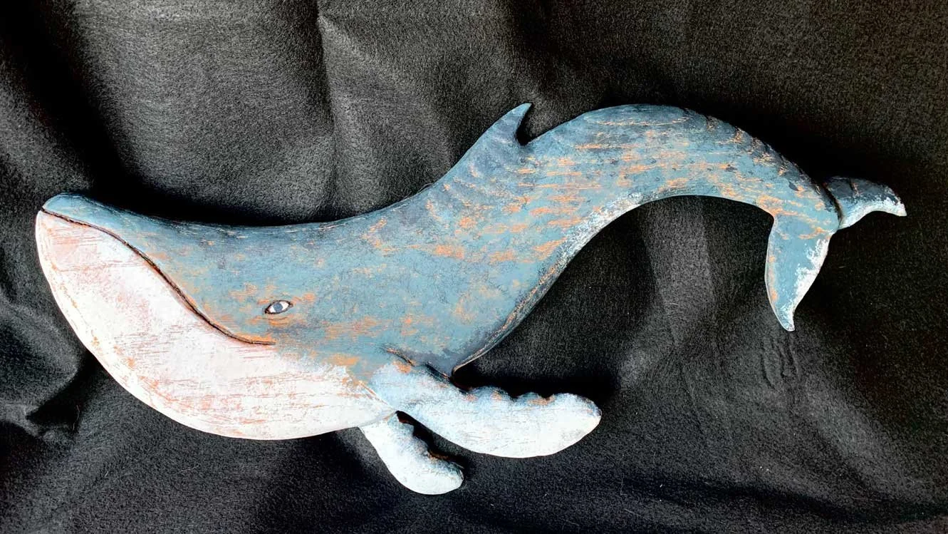 A decorative wooden whale sculpture painted in shades of blue, gray, and white, resting on a black fabric surface.