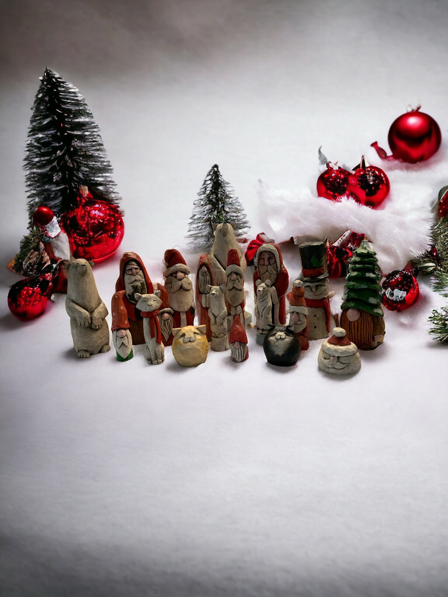 A Christmas scene with small figurines of Santa Claus, snowmen, and animals placed on snow-like surface. There are decorated Christmas trees, red ornaments, and faux snow in the background.