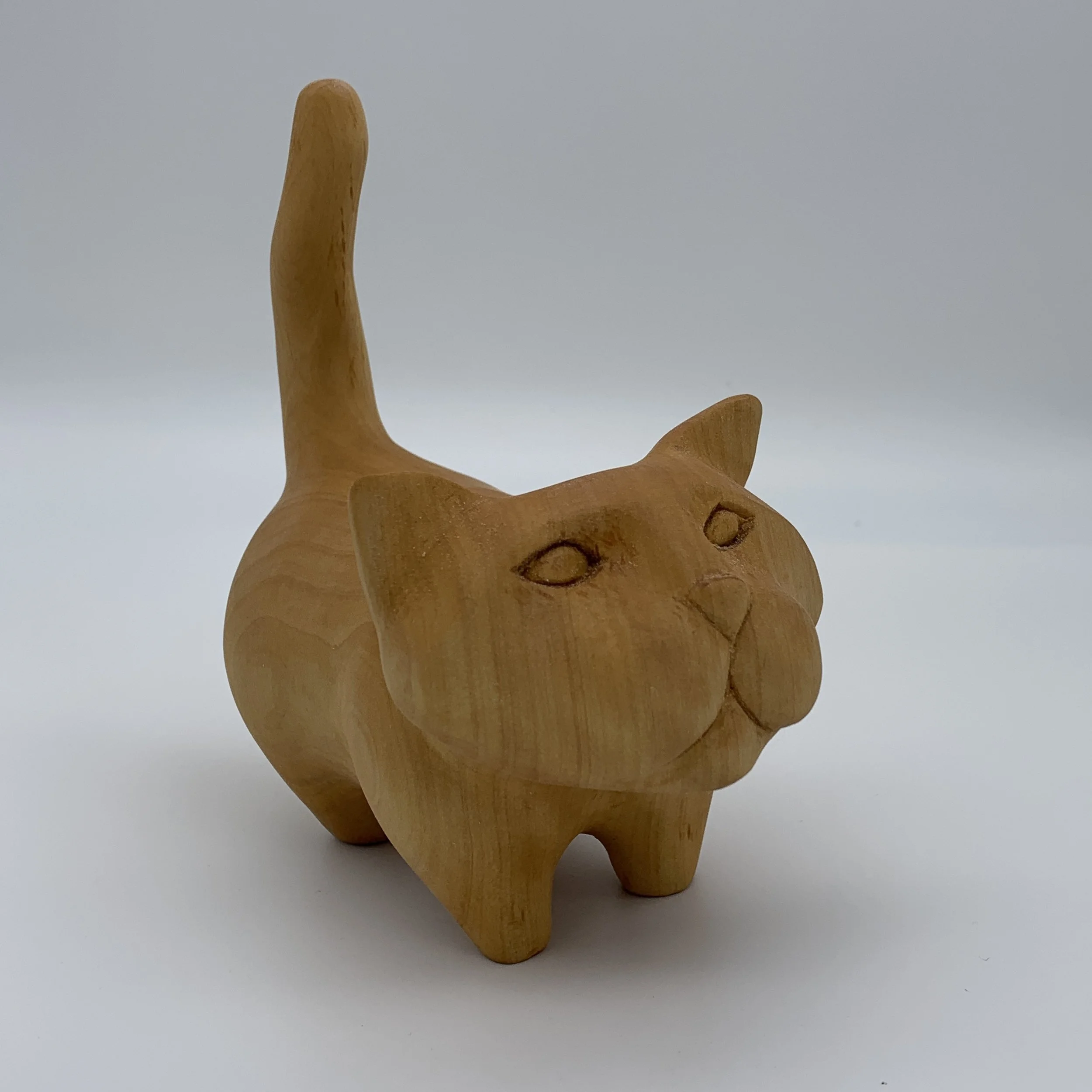 Wooden sculpture of a cat with a rounded body, pointed ears, and a long, curved tail, carved with minimal facial features.