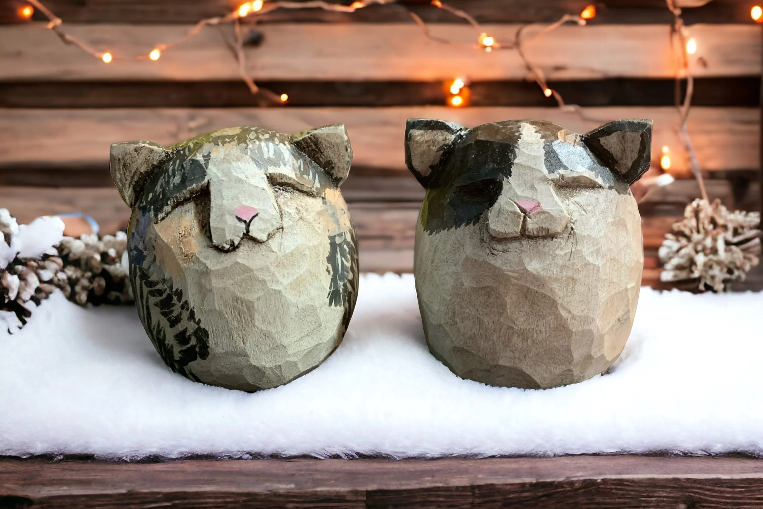 Two wooden animal figurines resembling cats, sitting on snow, with string lights and pinecones in the background.