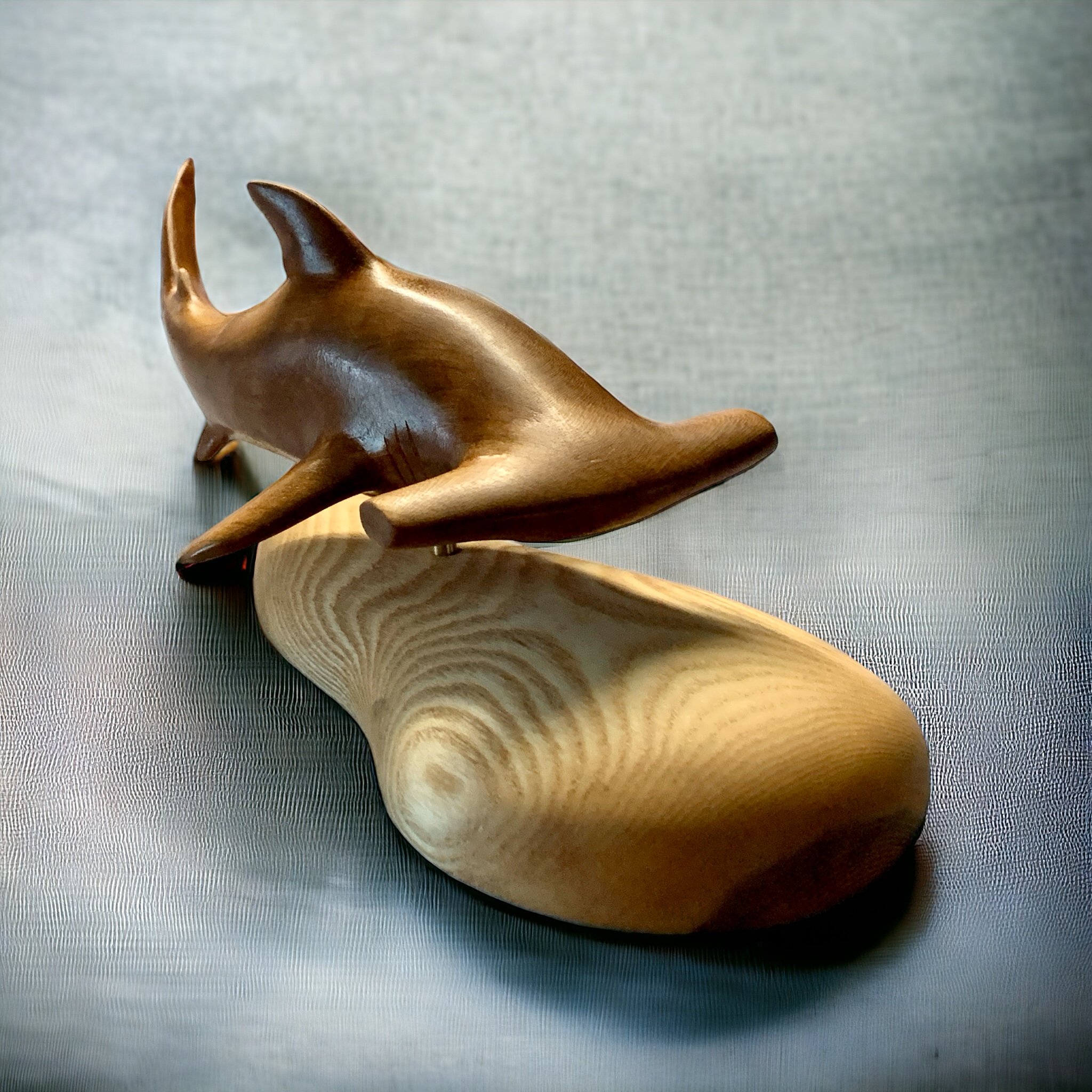 A handcrafted wooden sculpture of a hammerhead shark placed on a light-colored wooden stand against a gray background.