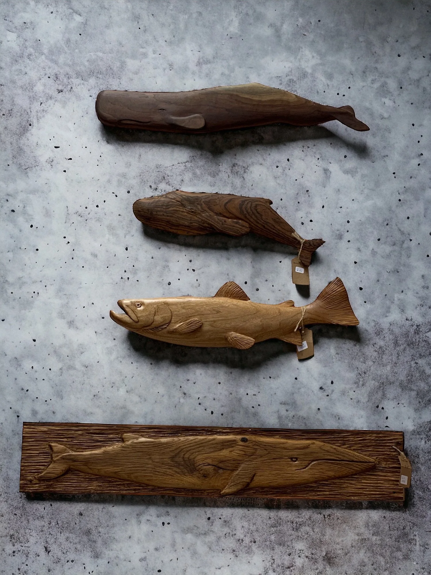 Four wooden sculptures of fish and whales arranged vertically on a gray, speckled surface. 