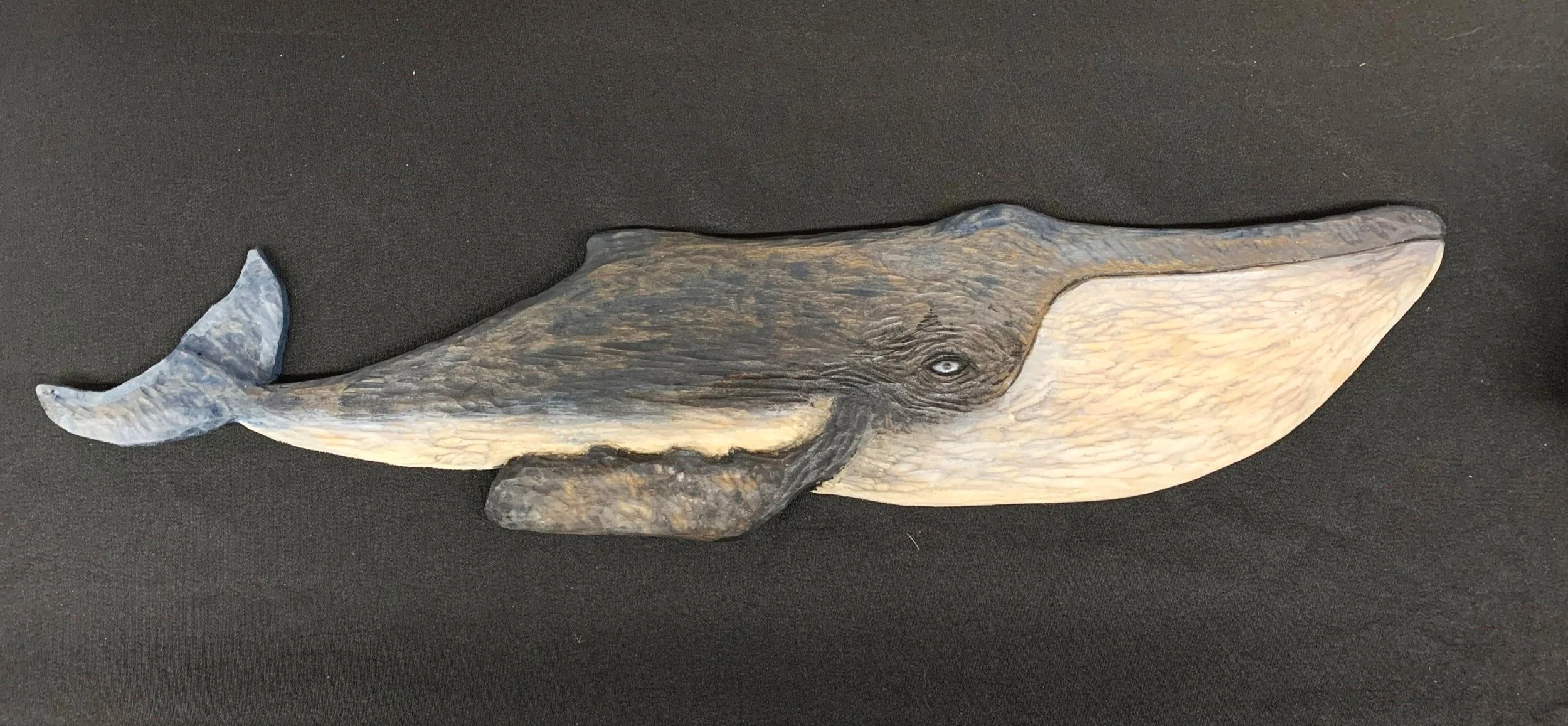 Wooden sculpture of a whale with detailed textures, dark and light shades, and a curled tail, placed on a black surface.