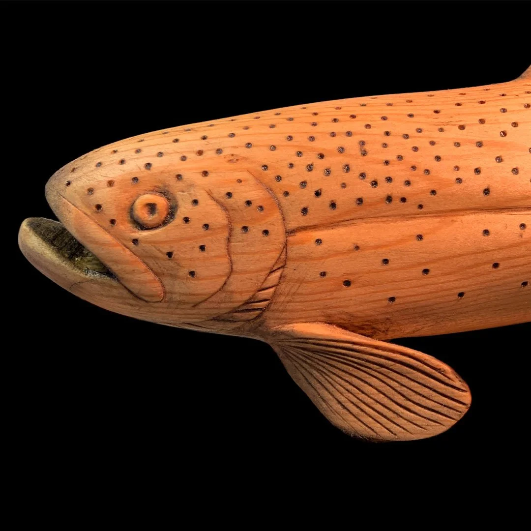 Close-up of a wooden fish sculpture carved to resemble a fish with detailed fins, a textured tail, and painted spots, against a black background.