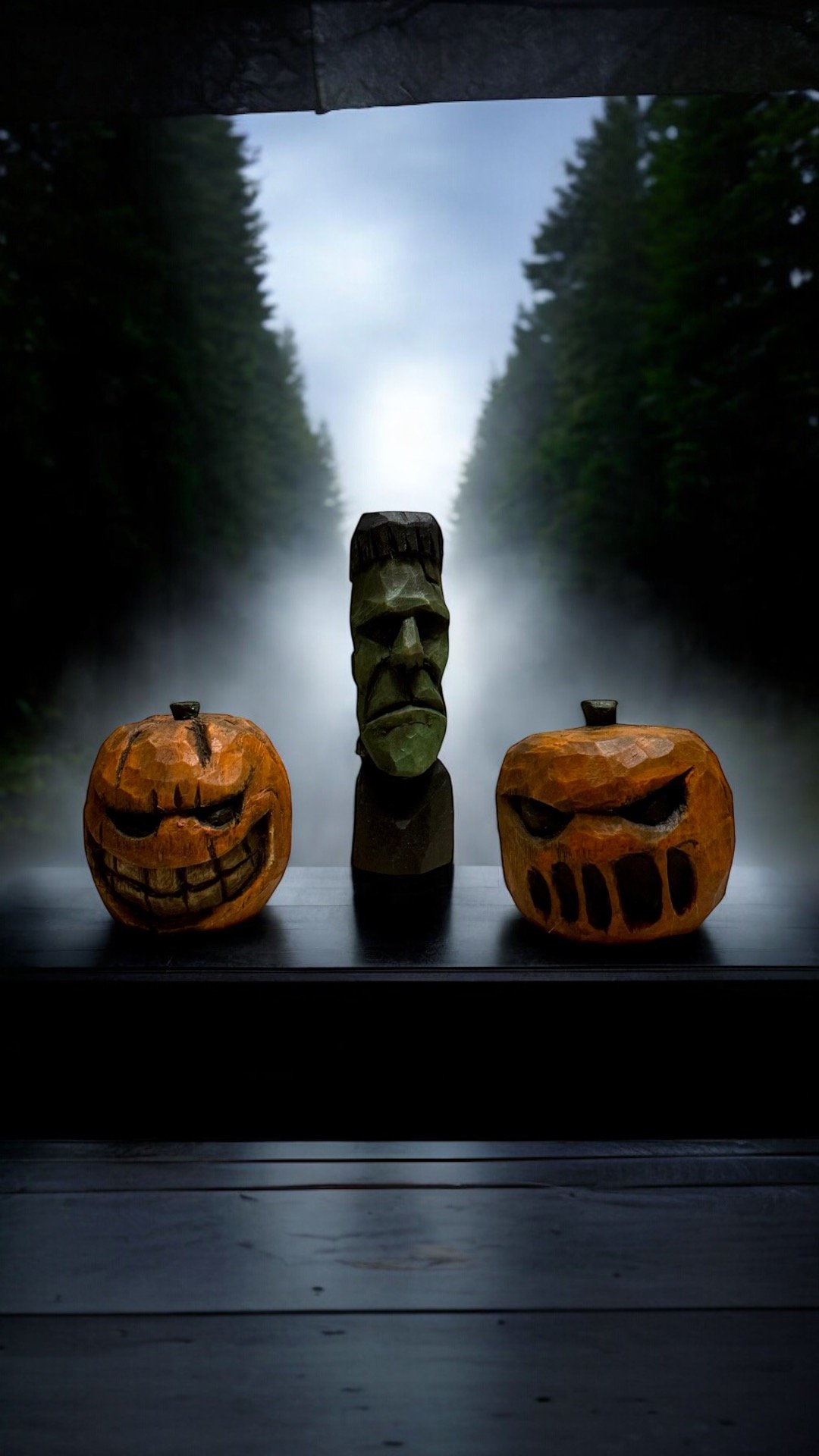 Three carved wooden Halloween decorations on a windowsill with a misty forest view in the background. The decorations include two jack-o'-lanterns with carved faces and a green-faced tiki-like figure in the center.