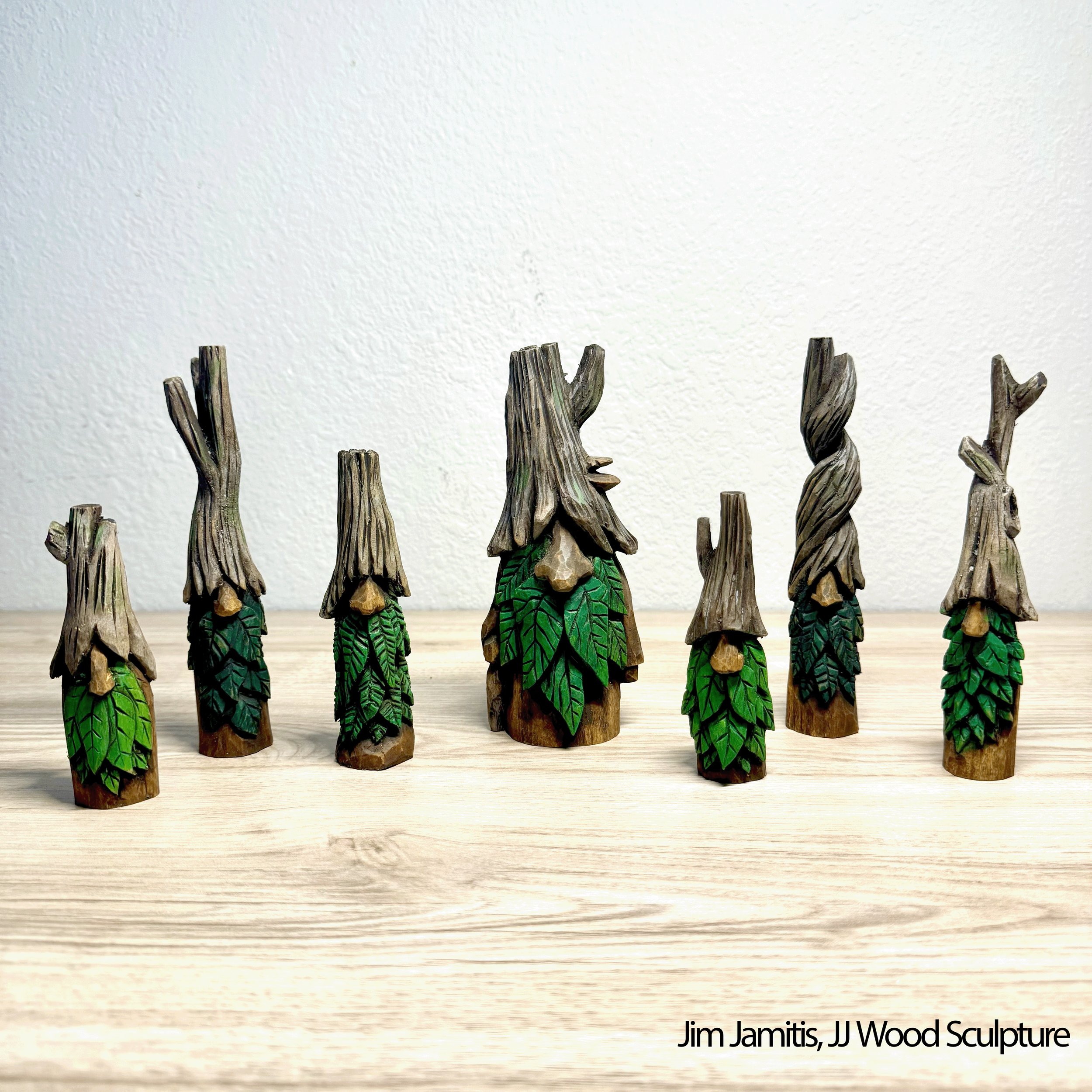 Seven carved wooden figures resembling tree stumps with faces, green painted leaves, and textured bark details, arranged in a row on a wooden surface.