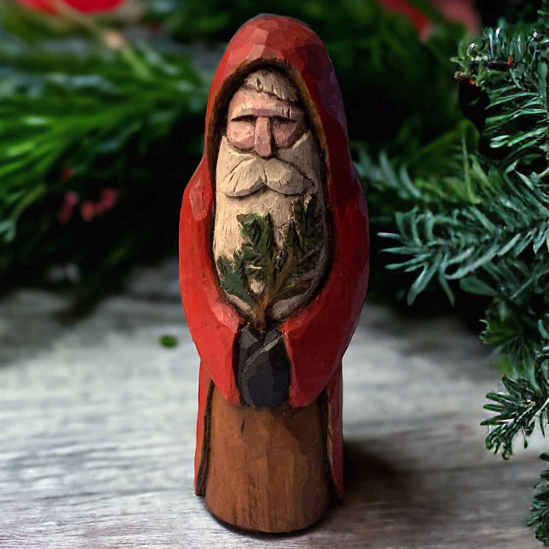 A wooden carved Santa Claus figure with a red coat, a white beard, holding a green pine branch, set against a background of Christmas greenery.