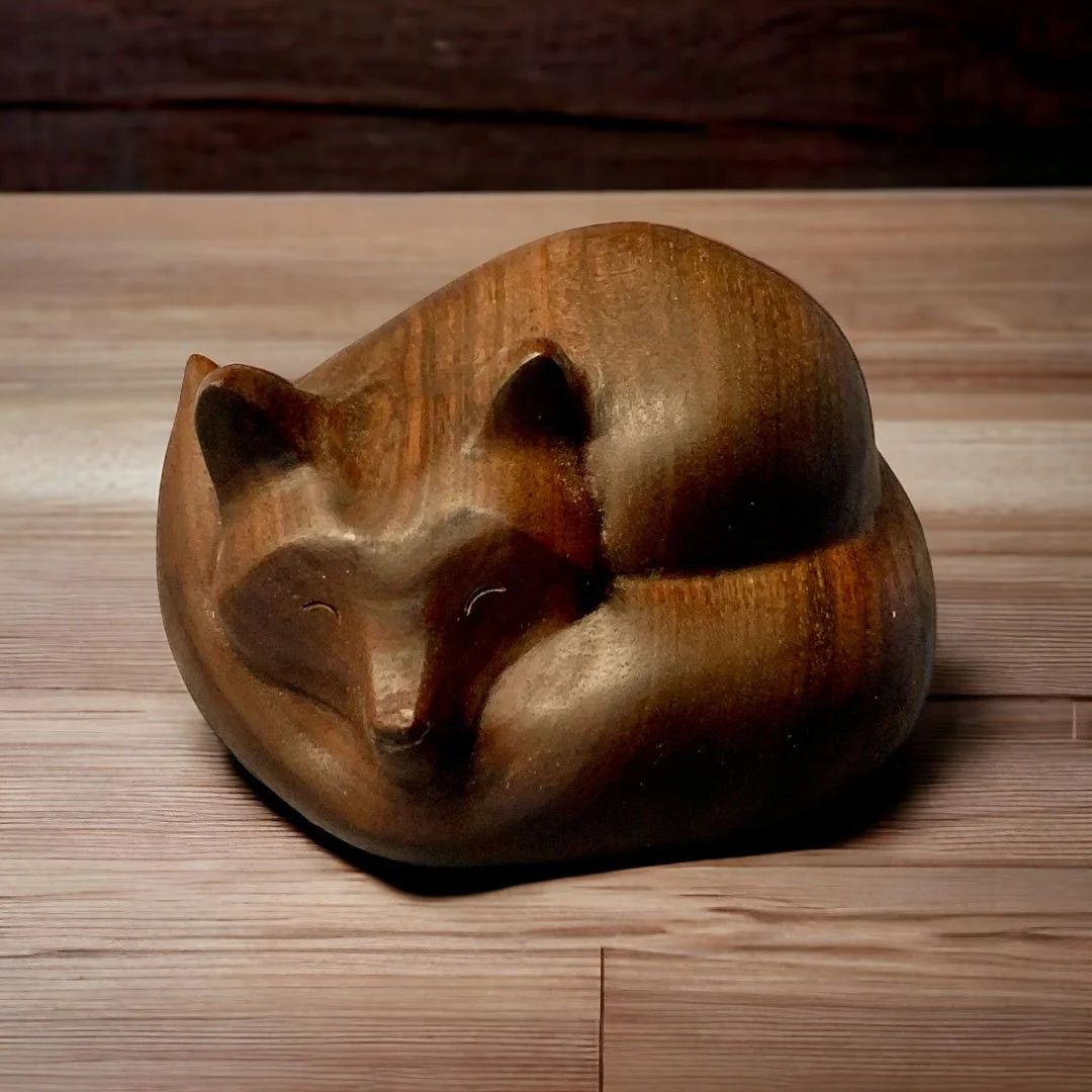 A wooden sculpture of a sleeping fox with its body curled up and eyes closed, placed on a wooden surface.