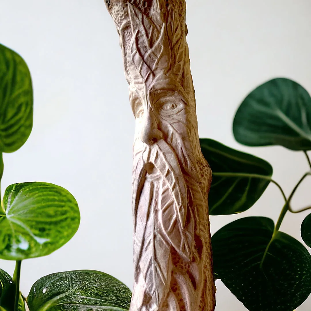 Wood carving sculpture of an elderly face with a long beard, surrounded by green leafy plants.