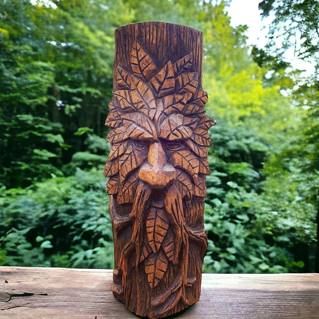 Wooden carved sculpture of a face with a beard and hair made of leaves, outdoors with green trees in the background.
