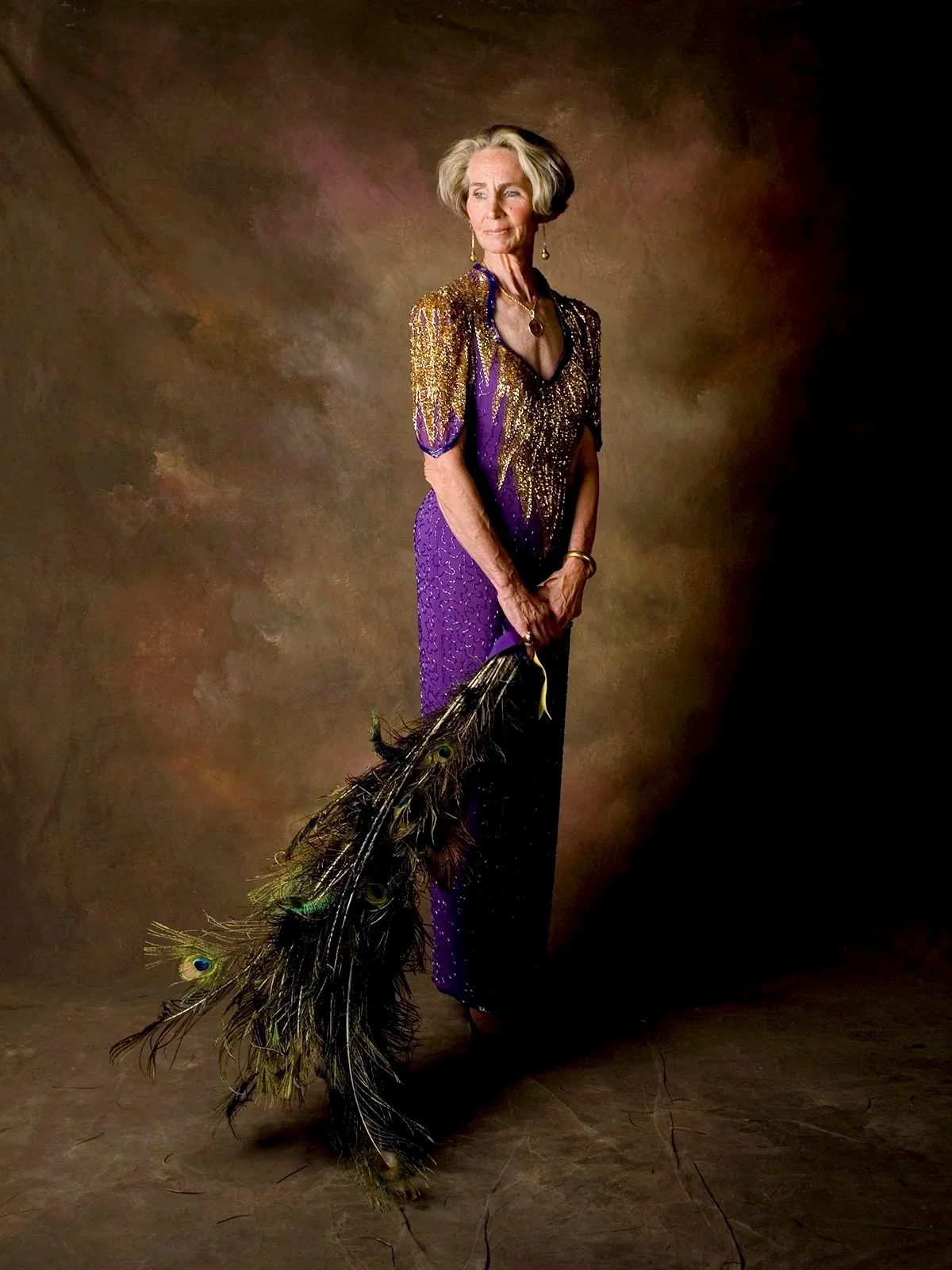 A tall woman wearing a purple and gold floor-length gown holds a handful of long, iridescent peacock feathers.