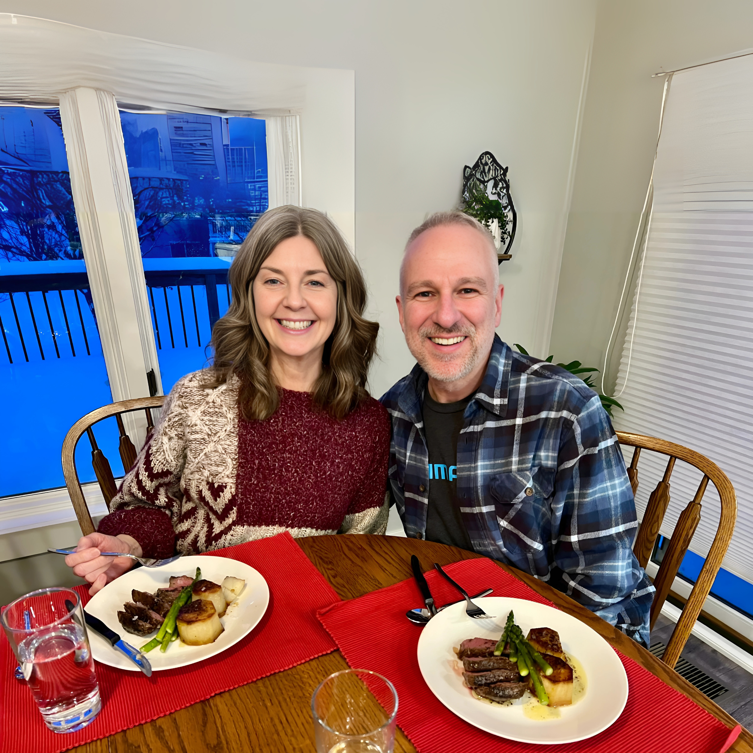 A smiling woman and man sitting at a dining table with plates of steak, asparagus, and potatoes, inside a well-lit room during dinner.