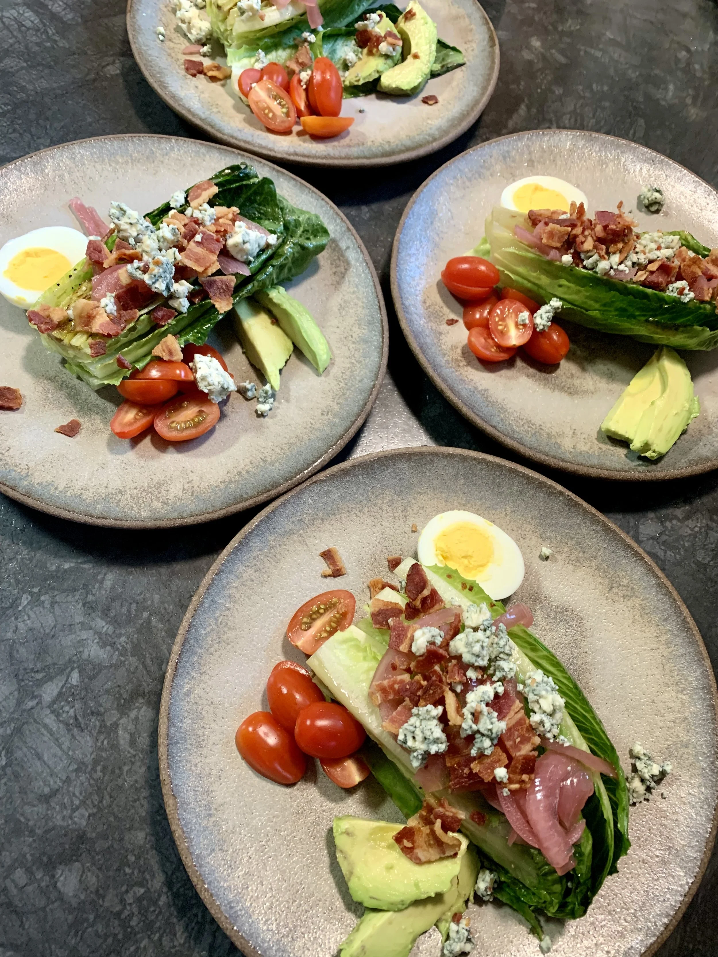 Four plates of chopped kale salad with cherry tomatoes, hard-boiled eggs, avocado, blue cheese, bacon, and red onion.