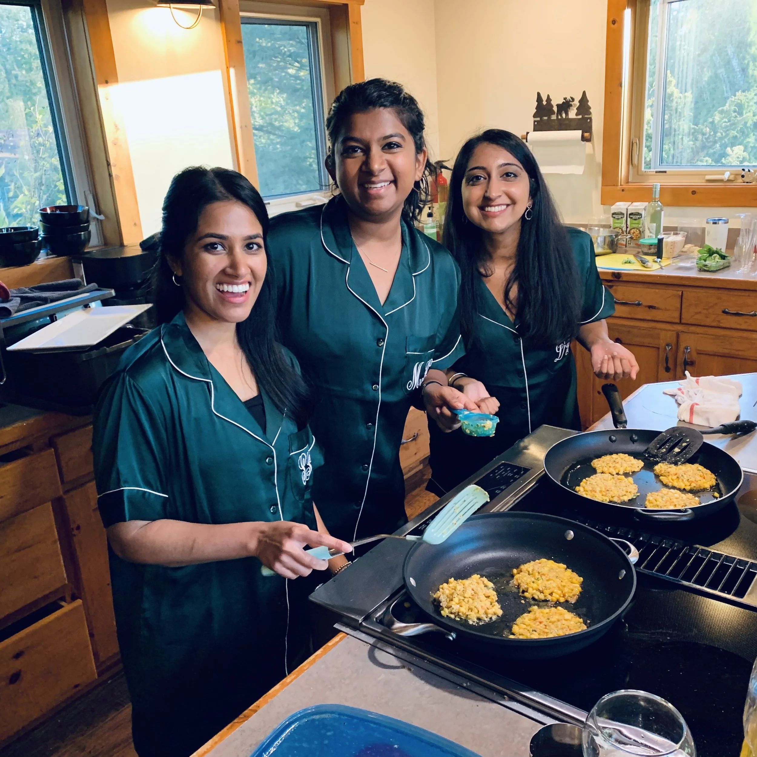 Three women in matching teal pajamas cooking breakfast in a kitchen with wooden cabinets and large windows.