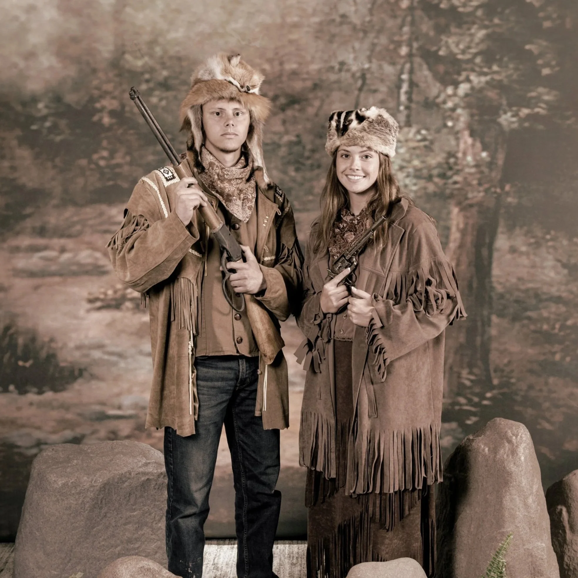 Two teenagers, a male and a female, dressed in western-style clothing, standing outdoors in front of a backdrop of trees. The male is wearing a fur-lined hat and holding a rifle, while the female is smiling, wearing a fringed jacket and holding a handgun.