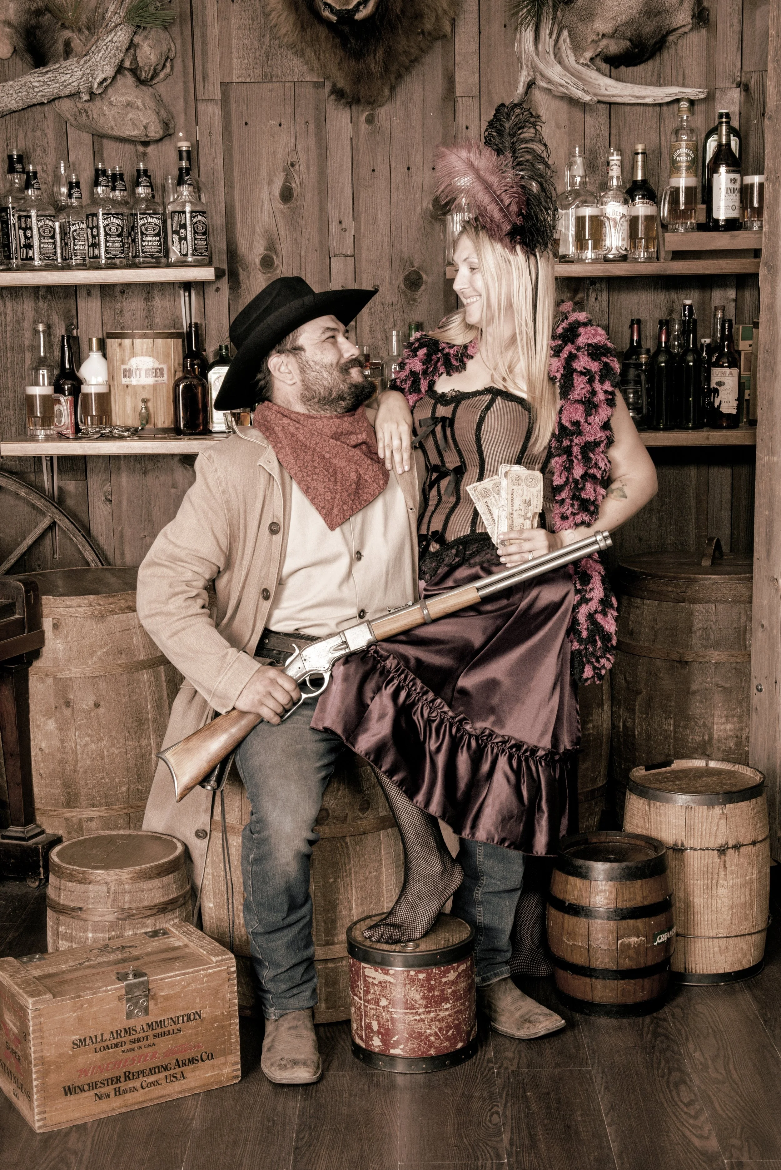 Scene with two people dressed in cowboy costumes; man with a black hat and beige coat holding a rifle, woman with a feathered headpiece and vintage dress holding cash, in front of wooden shelves with alcohol bottles, surrounded by barrels and vintage wooden boxes.