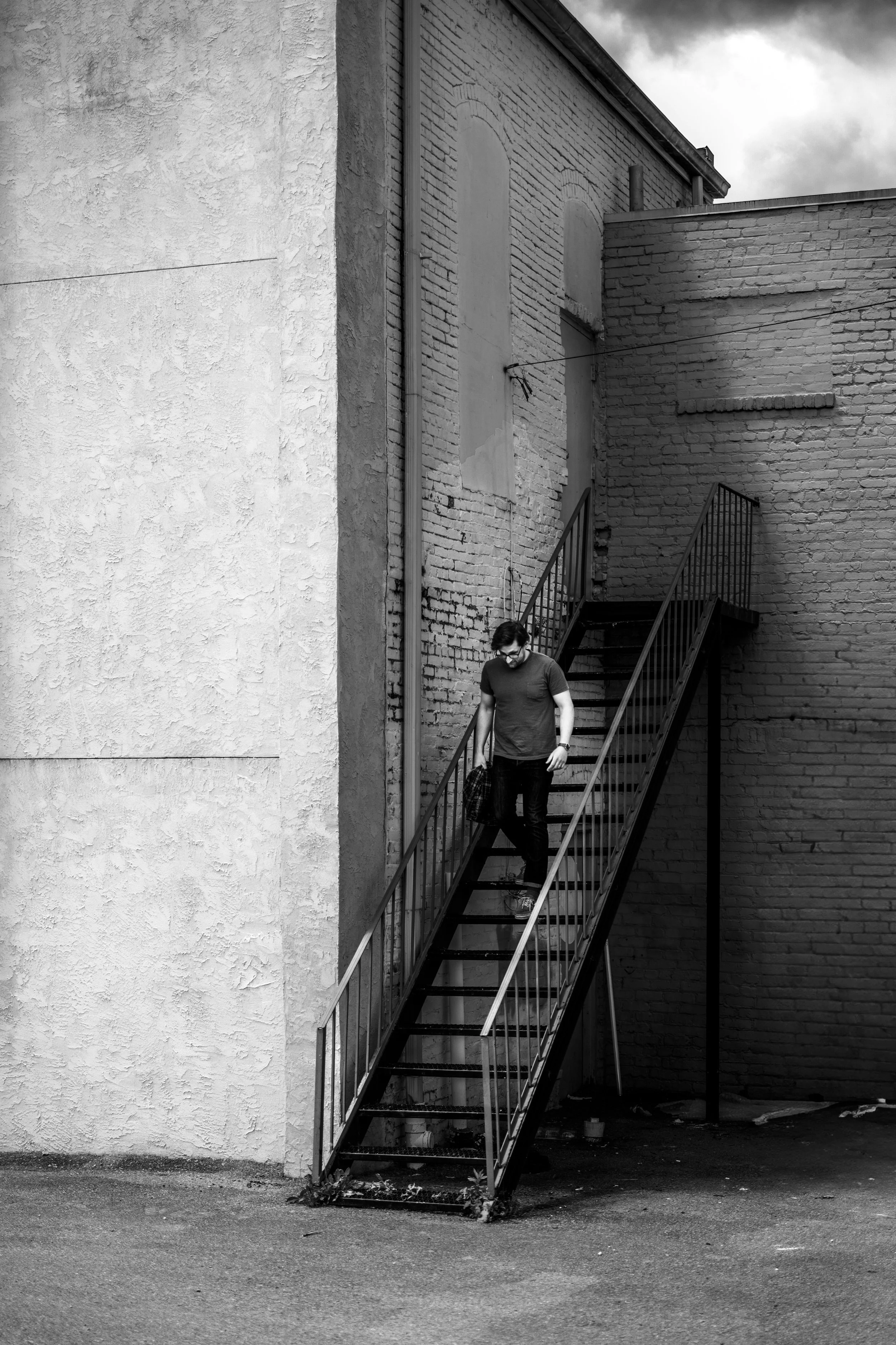 A person is walking down an outdoor metal staircase attached to a brick building, holding a bag, with a cloudy sky overhead.