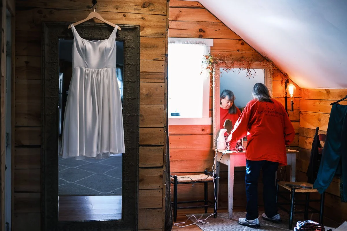 A wedding dress hanging on a mirror in a wooden room, with a person in a red shirt sitting at a vanity mirror with a mirror, surrounded by chairs and personal items.