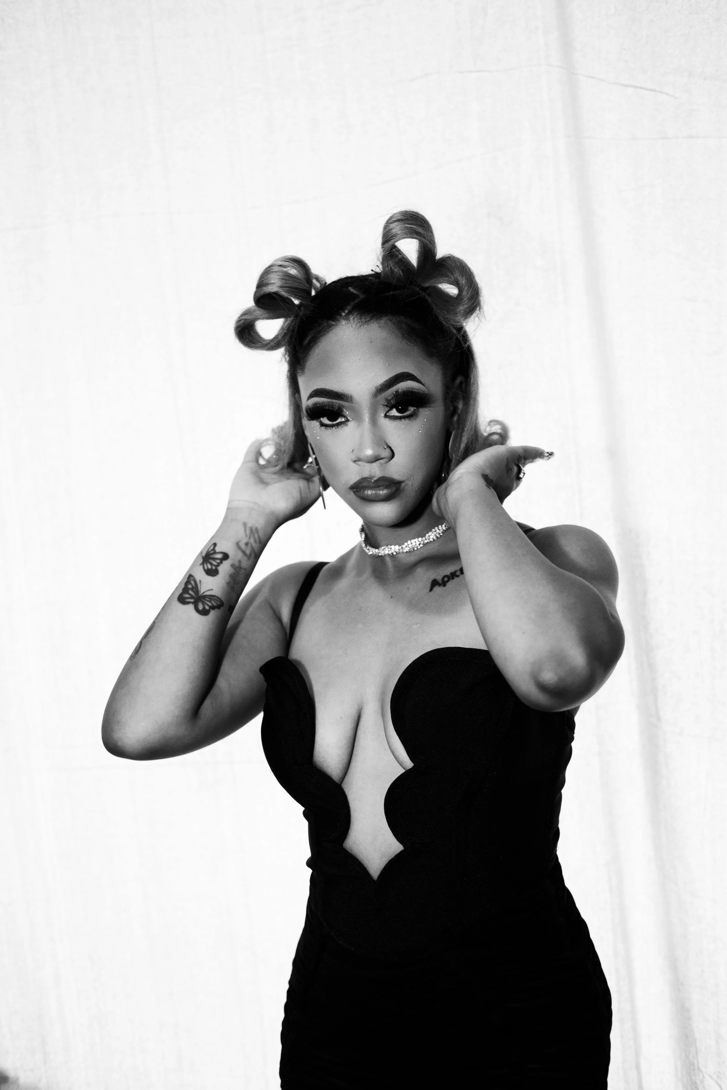 A woman with styled hair in buns, bold makeup, a necklace, tattoos, and a revealing black dress poses in front of a light background.