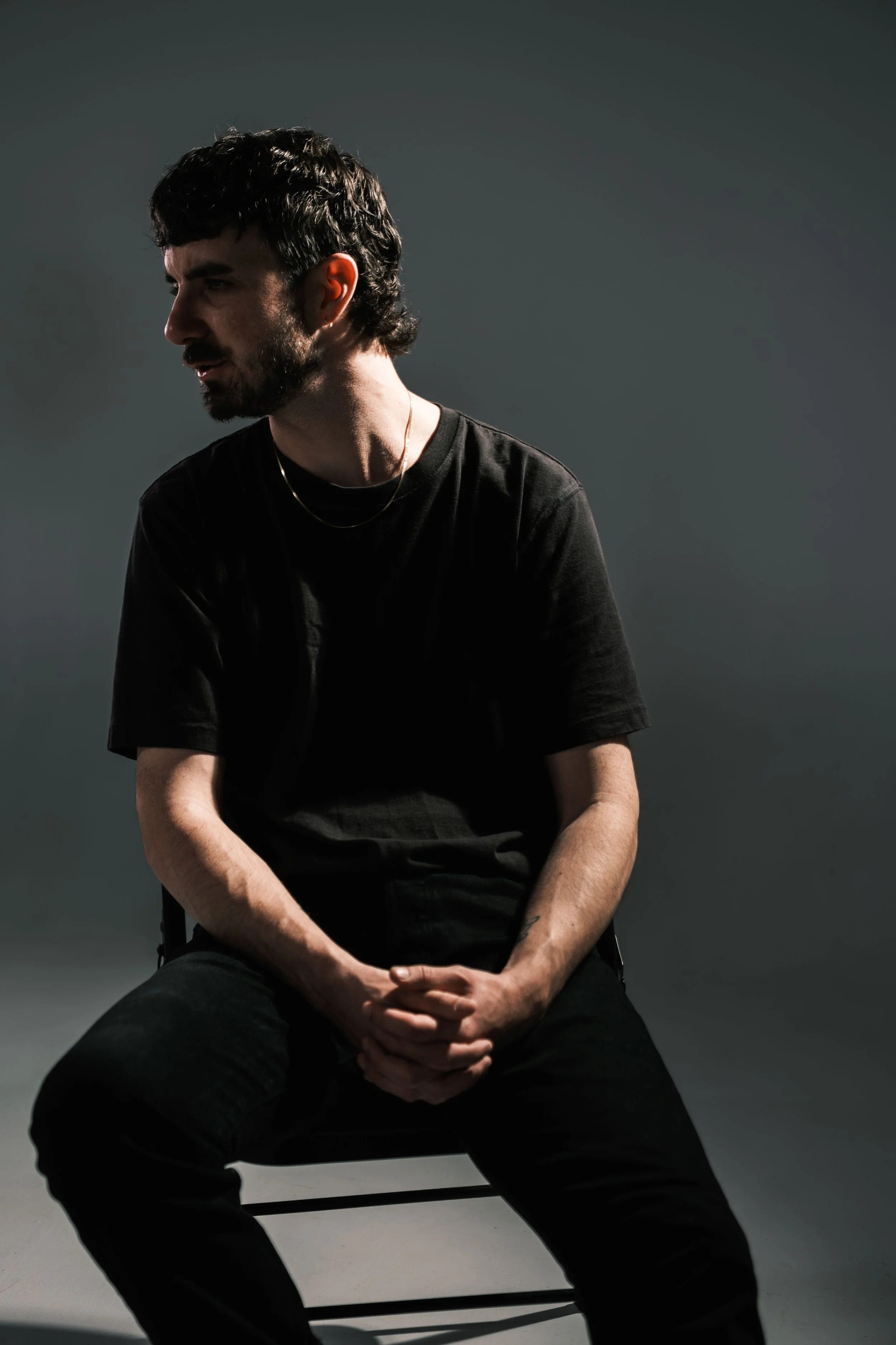 A man with dark hair and a beard, wearing a black t-shirt and gold chains, sitting on a stool against a dark background, looking to the side.