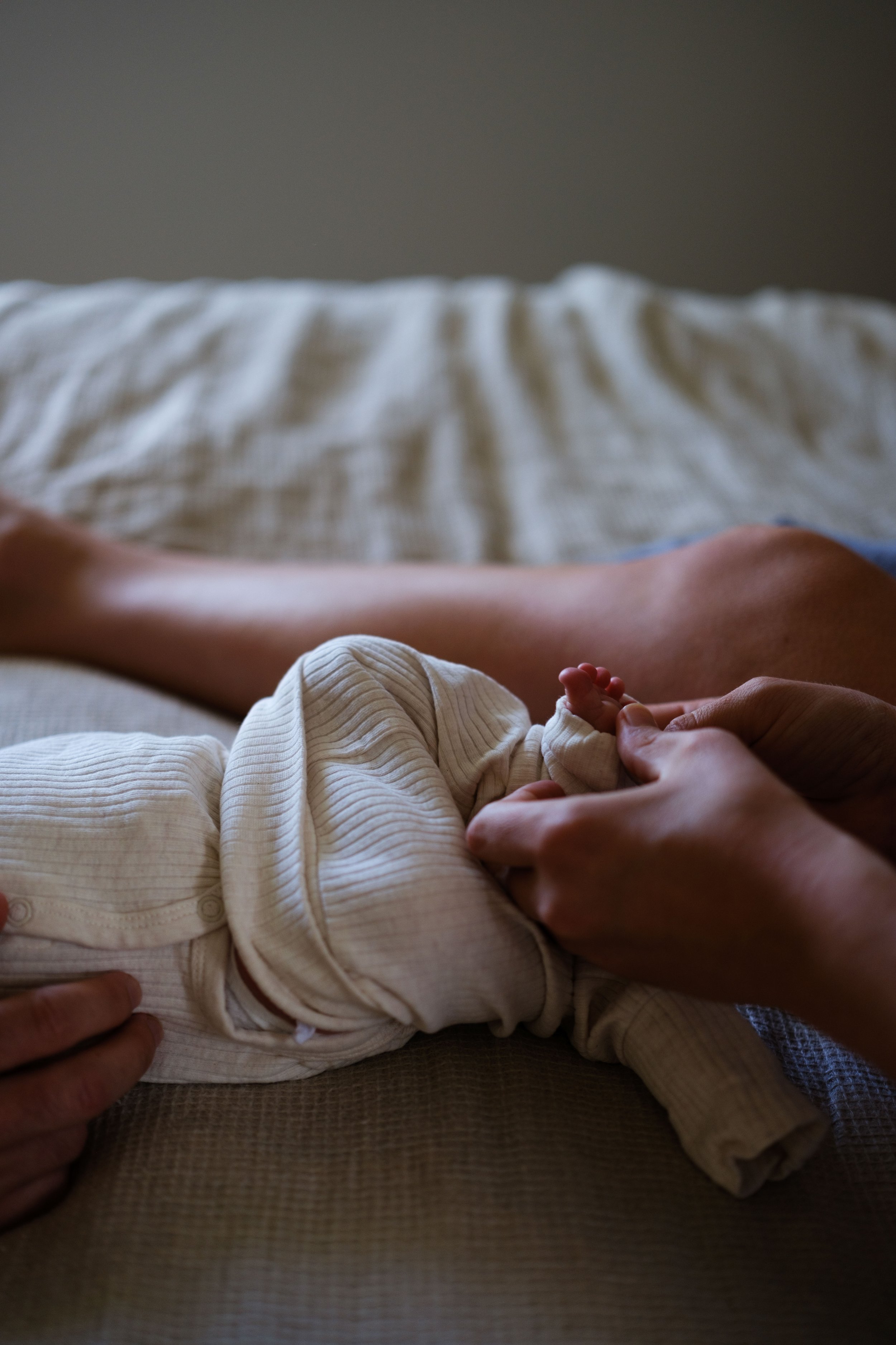Person holding a baby's hand, lying on a bed with beige sheets.