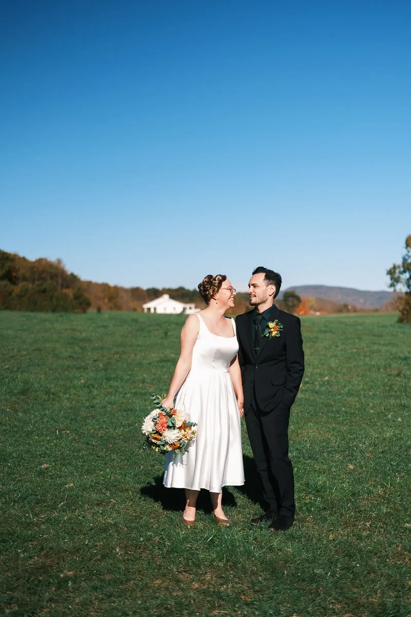 A couple in wedding attire standing on a grassy field outdoors, with a woman holding a bouquet of flowers and a man in a black suit, smiling at each other against a clear blue sky.