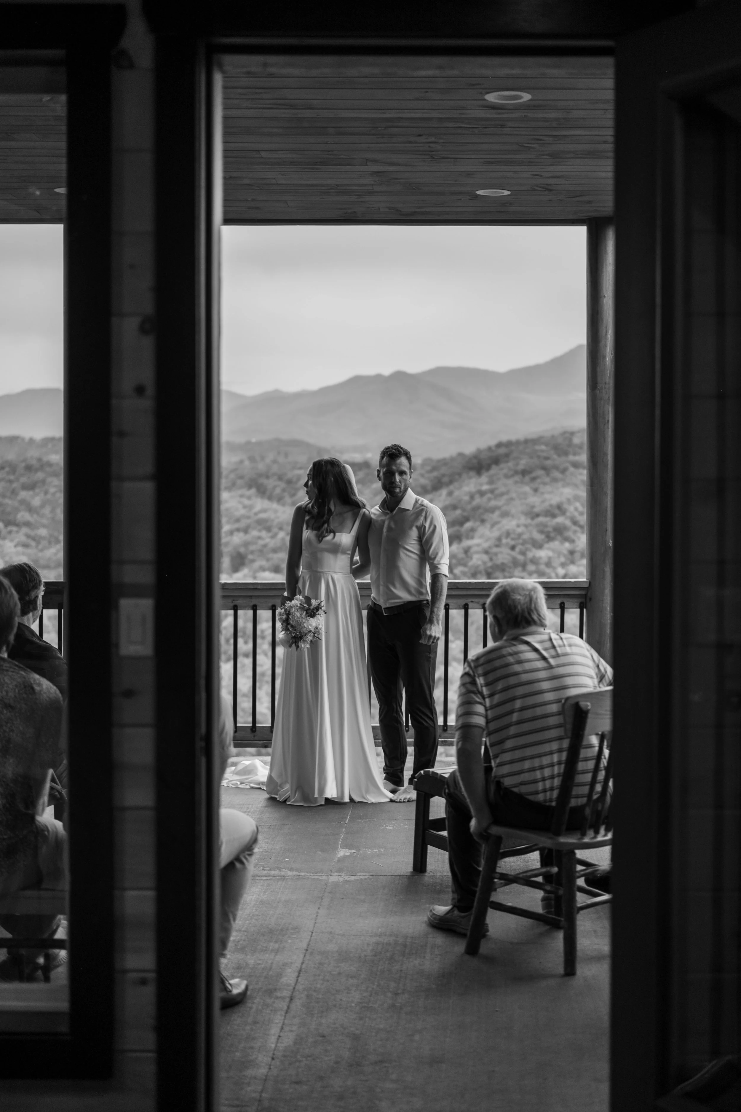 A black and white photo of a bride and groom standing on a balcony during their wedding ceremony, with scenic mountains in the background, and guests seated and observing.