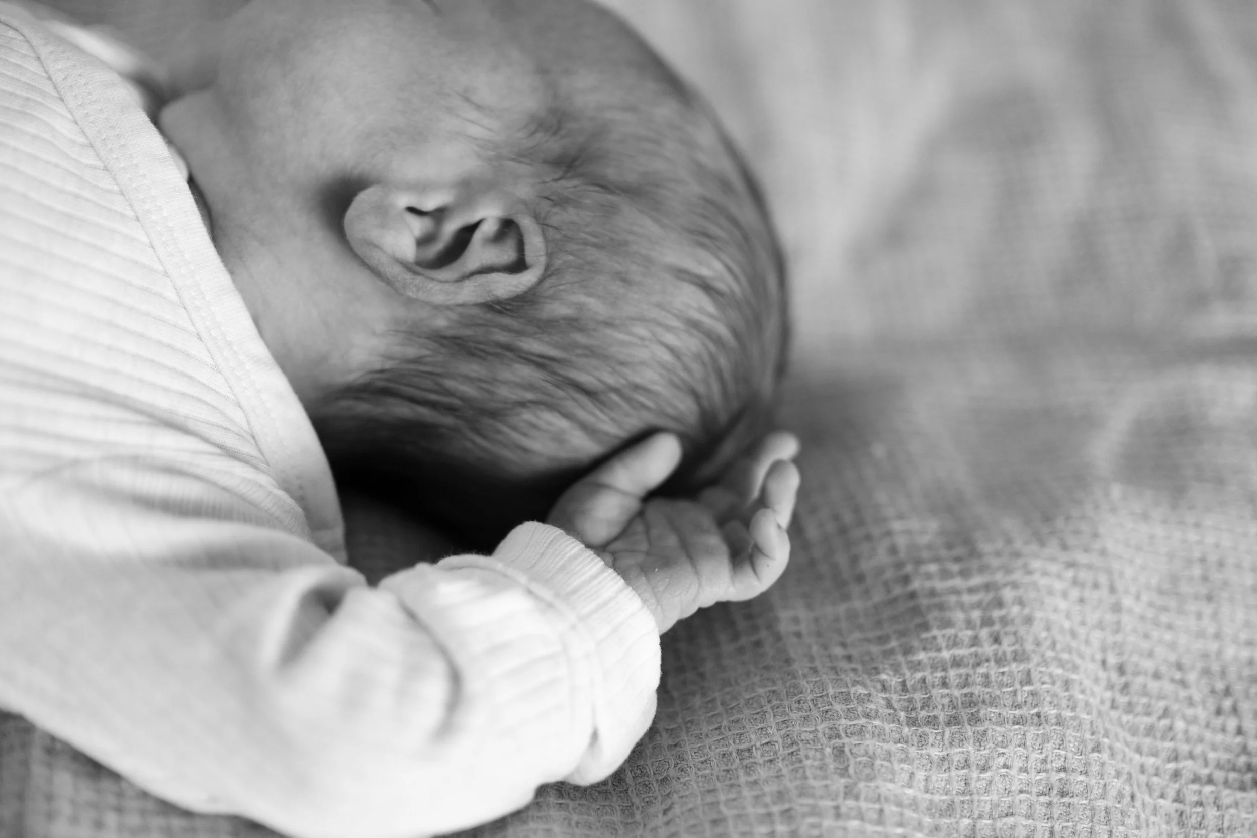 A black and white photo of a sleeping newborn baby lying on a blanket, with hand near face, view of head and ear.