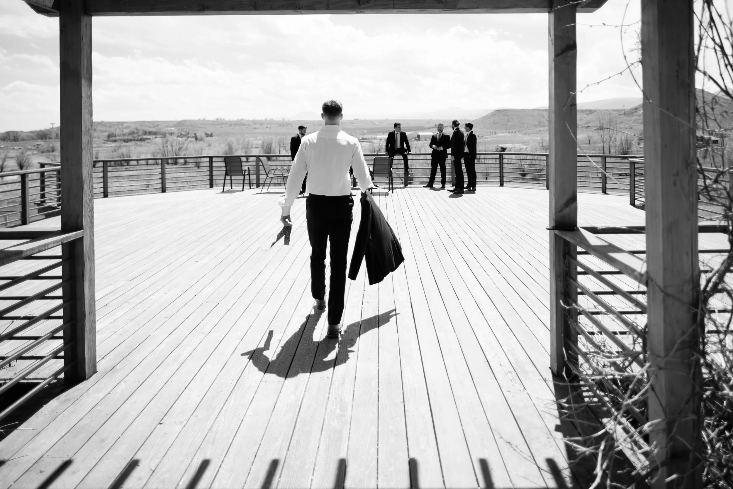 A man walking towards a group of five men on a wooden deck with a view of open countryside in the background.