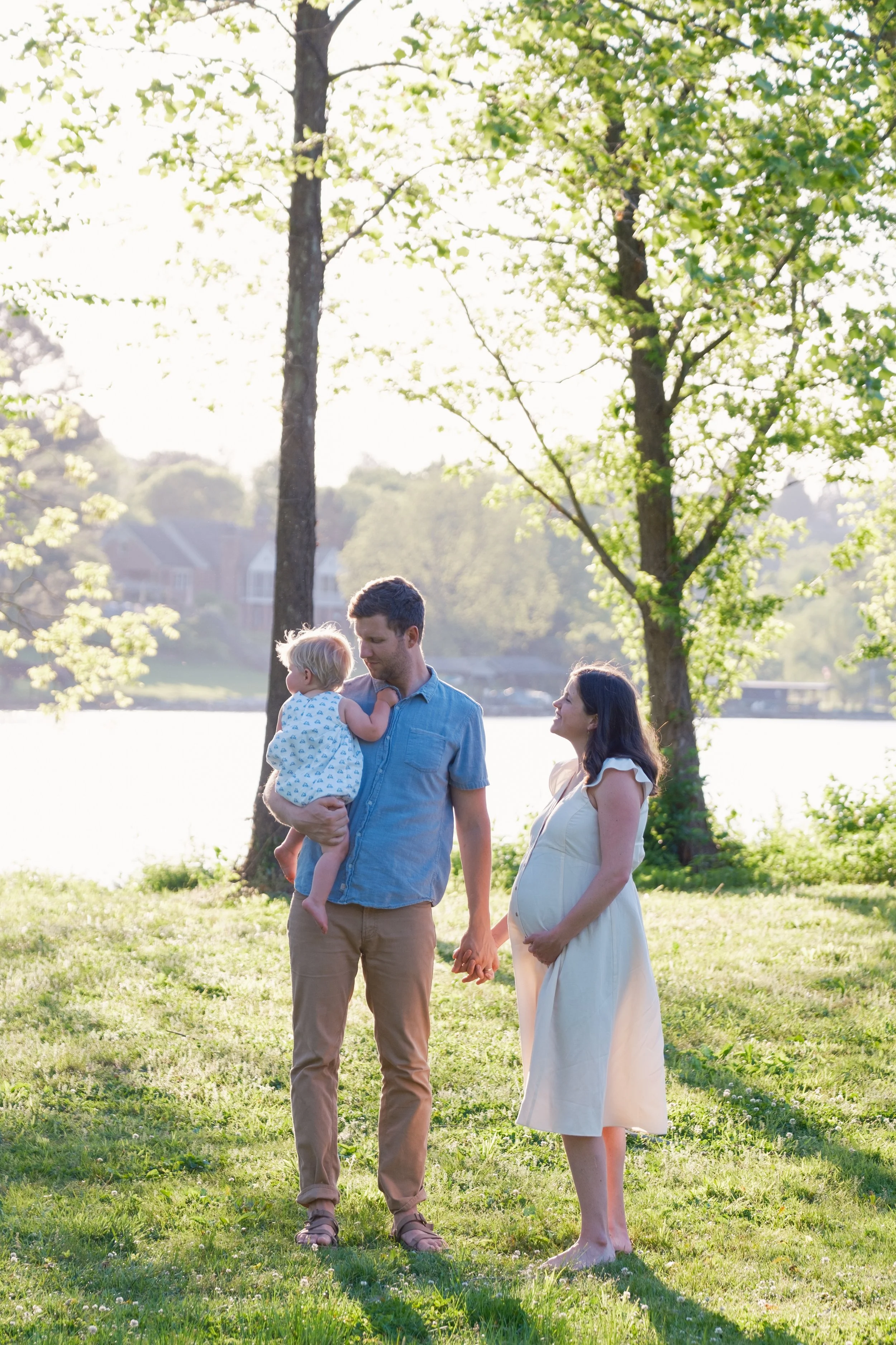 A pregnant woman and a man holding a child by the lake during sunset.