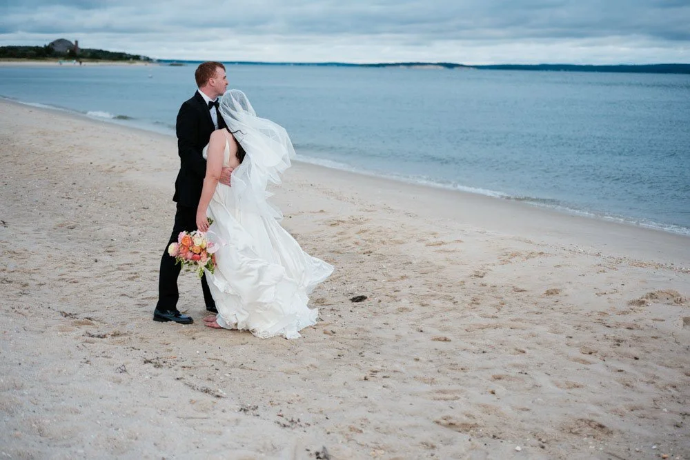 Groom holding bride in wedding dress on sandy beach with ocean in the background.