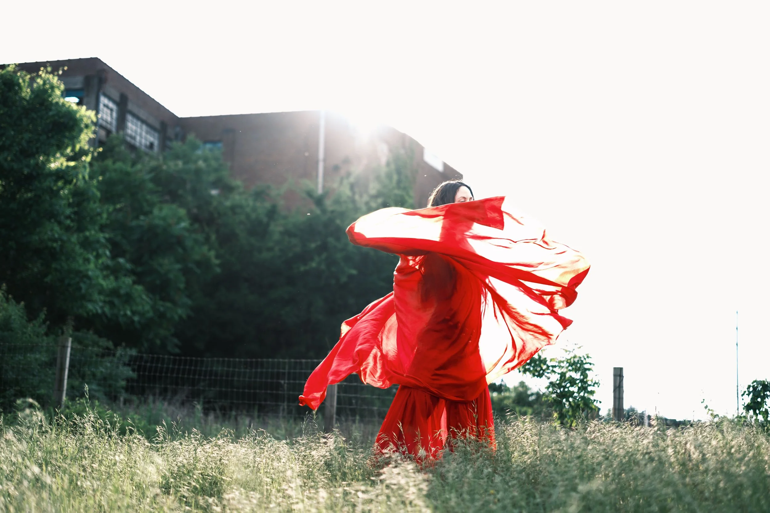 A woman in a flowing red dress twirls outdoors in a grassy field with trees and a building in the background, backlit by bright sunlight.